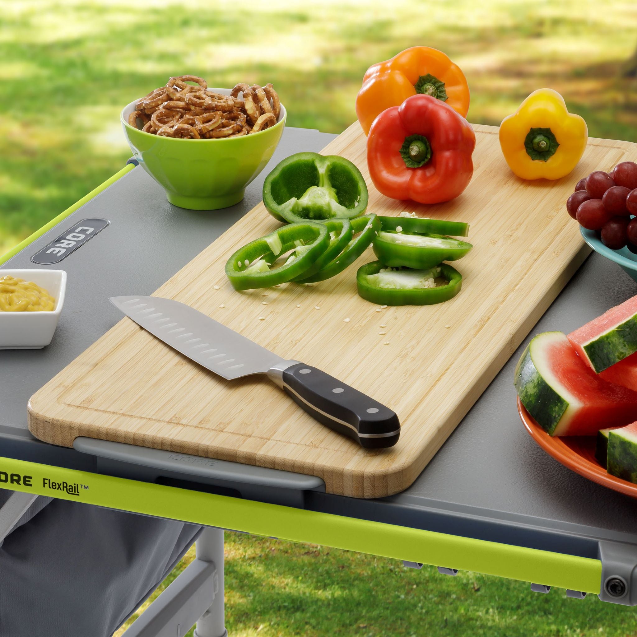 Outdoor table with cutting board, knife, and fresh vegetables on a grassy background.