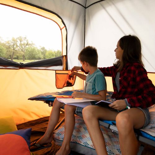 Two people are sitting inside a tent, with one reading a book and the other putting pencils in a storage pocket.