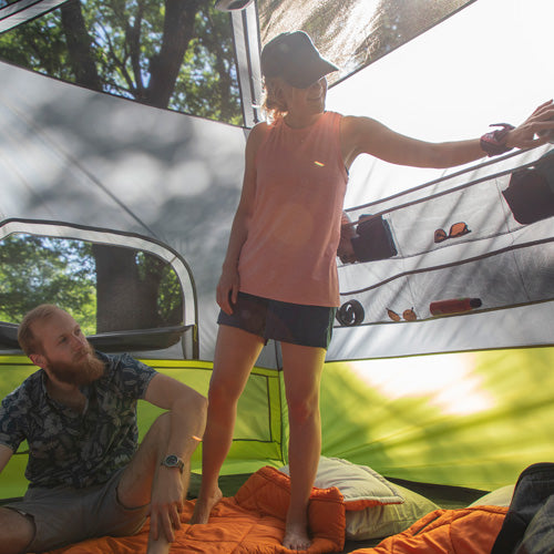 A person is standing inside a tent, reaching into the storage pockets, while another person is sitting.