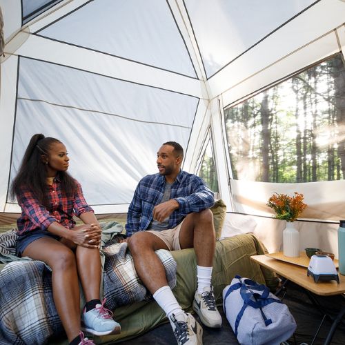 Two people sitting inside a CORE 6 person lighted instant pyramid tent, looking at each other. Sunlight streams through the tent's mesh windows, illuminating the surrounding forest.
