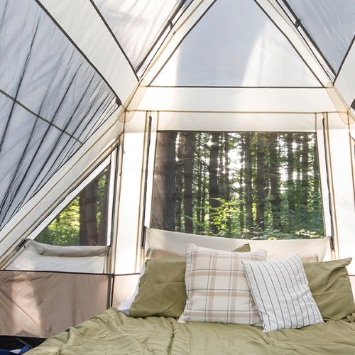 Interior of a 6 person lighted instant pyramid tent with a bed made with green bedding and pillows, looking out through a large window at a forest with tall trees.