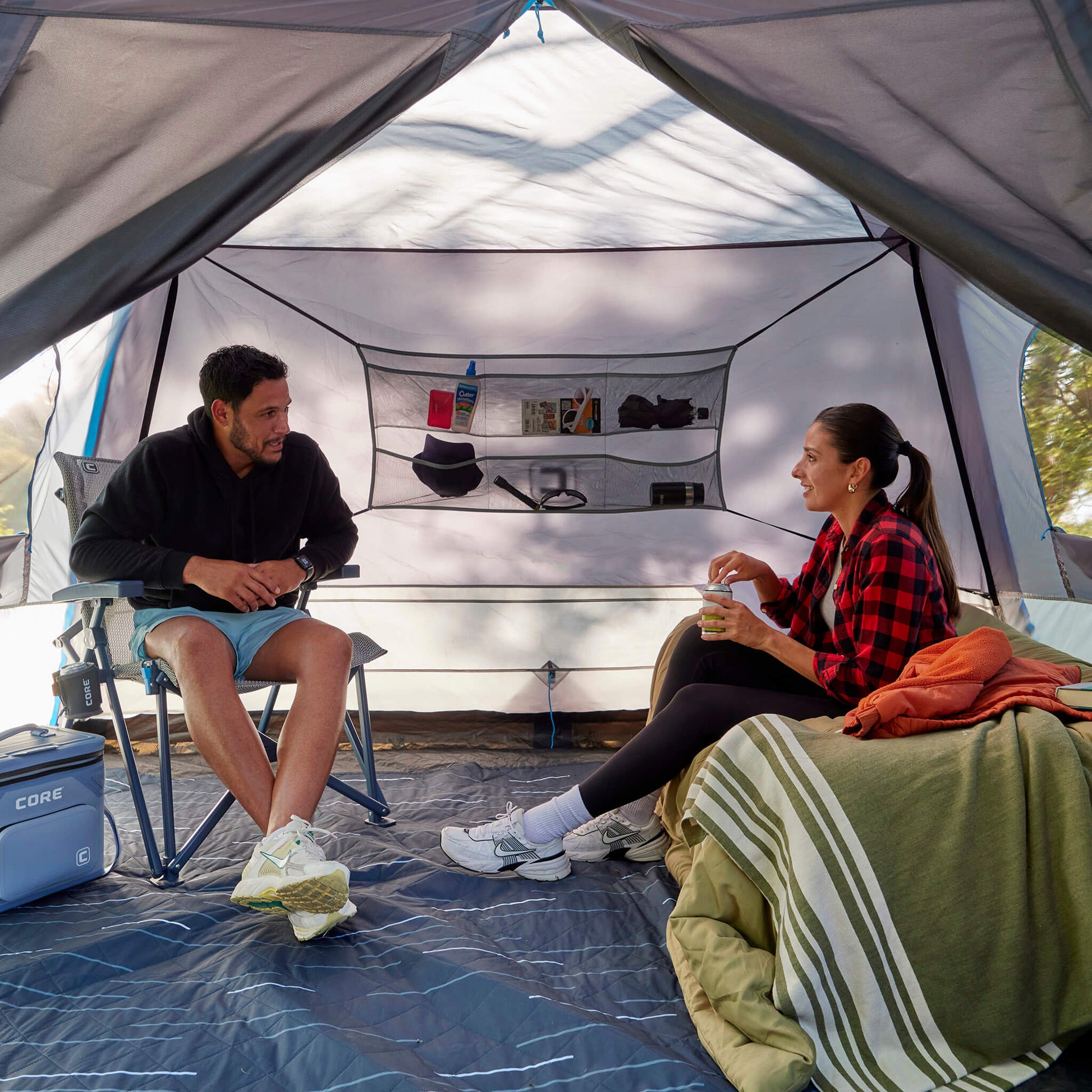 Friends sitting on a CORE chair and cot, enjoying time inside the CORE 6 lighted person instant cabin tent with full rainfly.