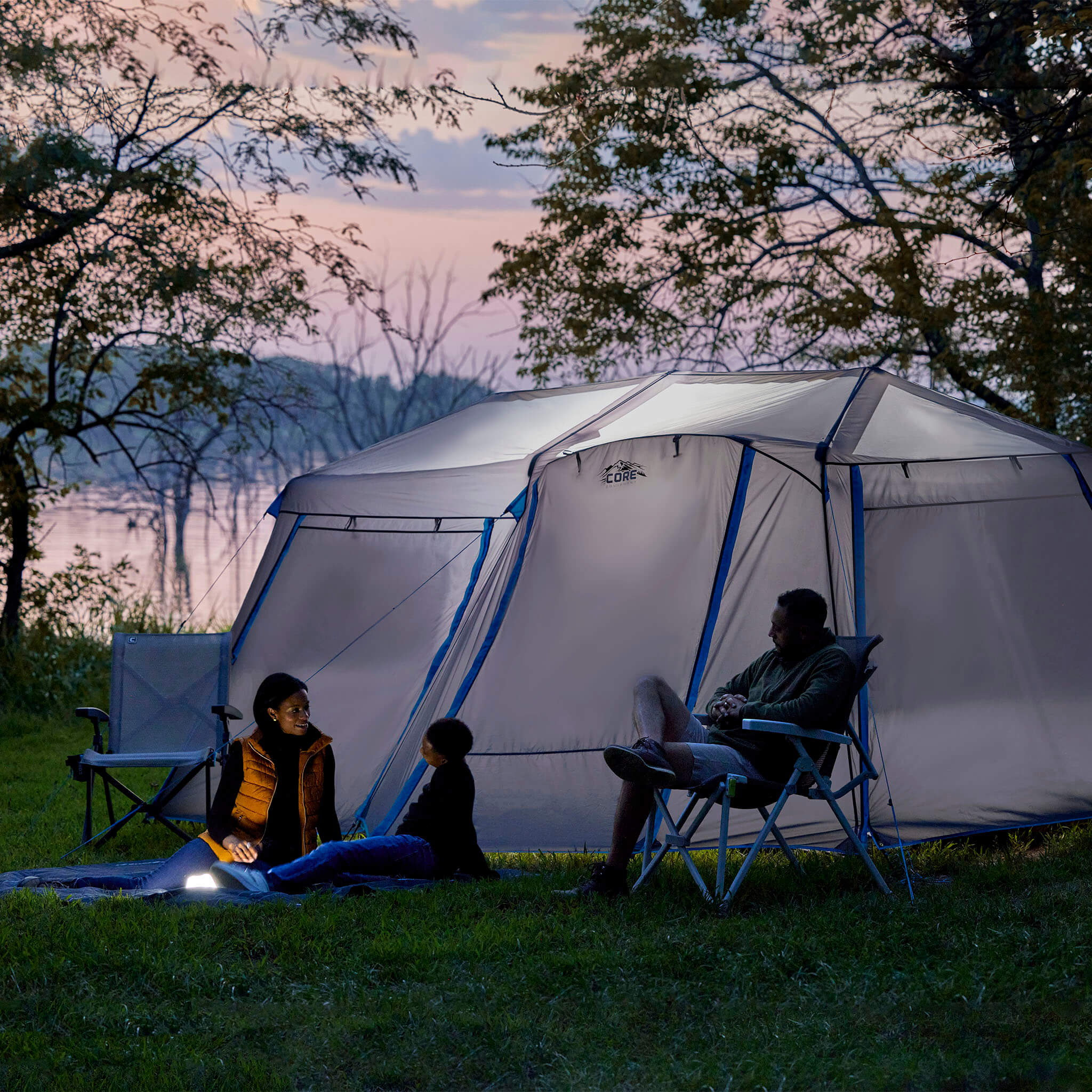 Family relaxing beside the CORE 9 person lighted instant cabin tent with full rainfly in blue and gray, designed for fast setup and reliable weather protection.