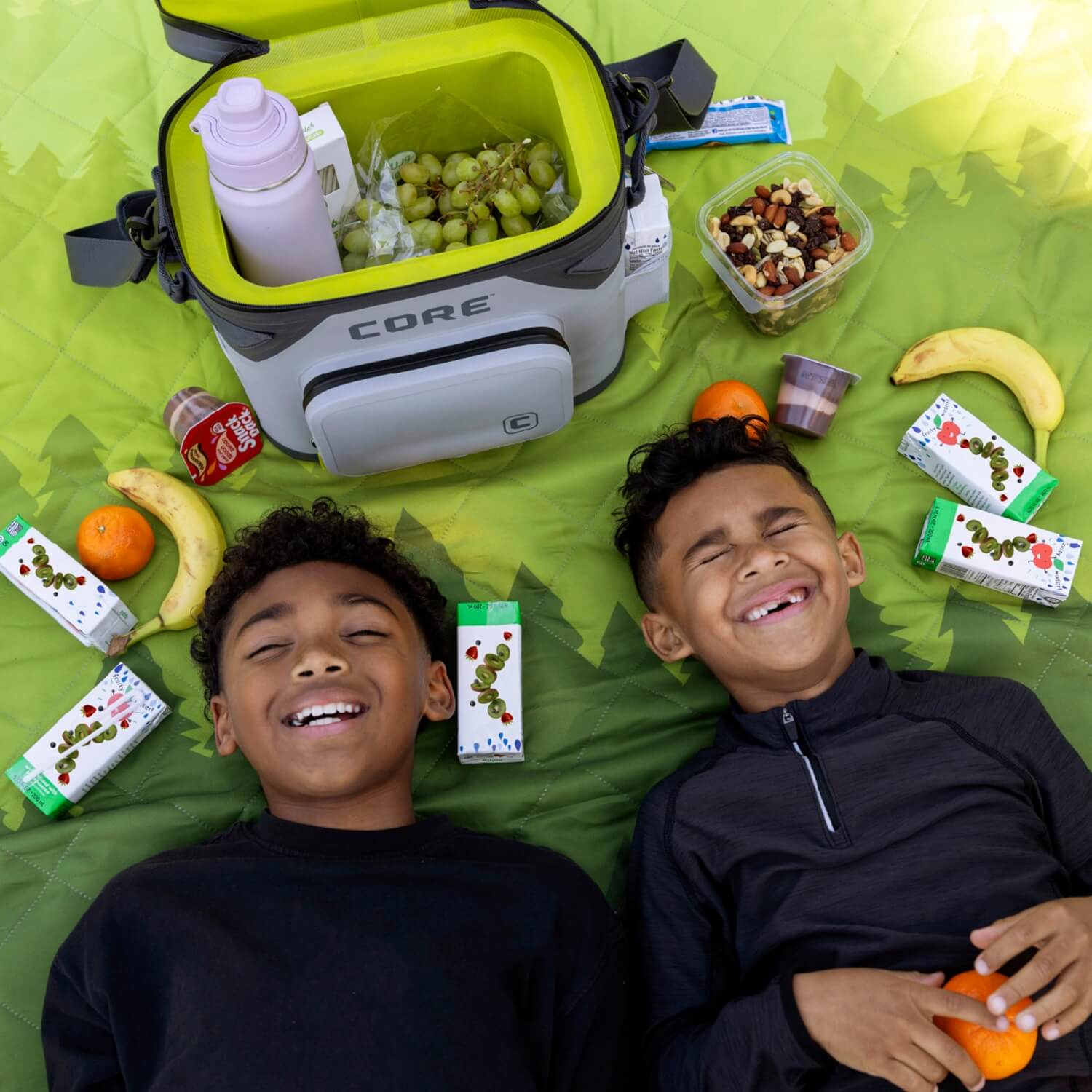 Two smiling children lying on a CORE green weighted outdoor blanket next to an open CORE Climakeep soft cooler filled with grapes and snacks. Around them are juice boxes, bananas, oranges, and trail mix, suggesting a fun picnic setup.