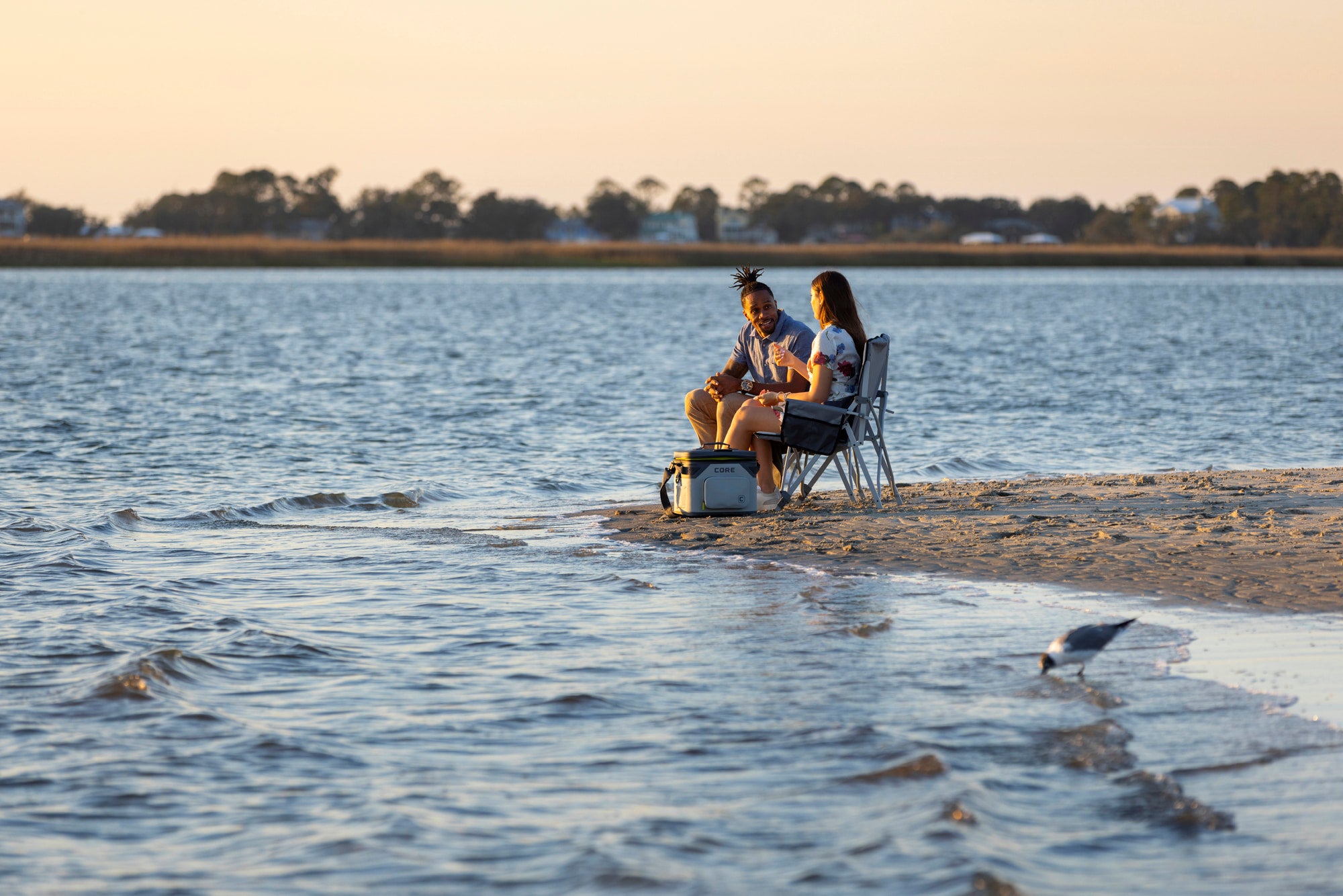 Two people sitting on chairs at the beach by the water at sunset with CORE Climakeep 18 can soft cooler next to them.