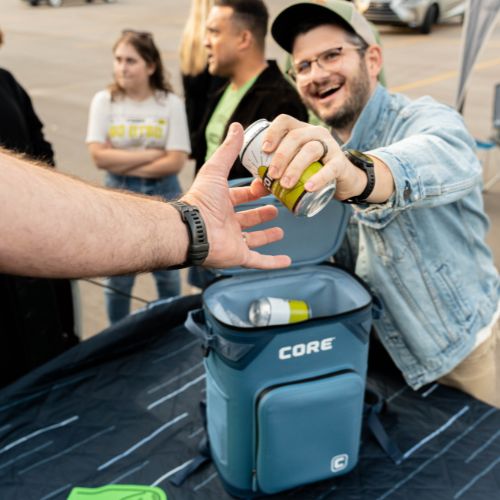 A person opening a Navy 'CORE' backpack cooler with drinks inside, surrounded by people in an outdoor setting.