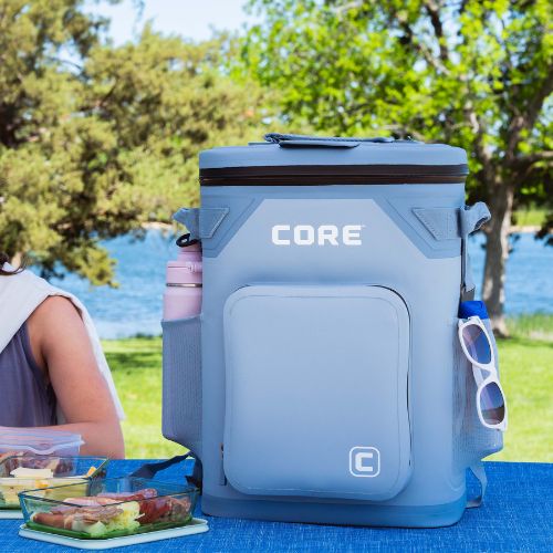 A blue 24 can backpack cooler with 'CORE' branding on a picnic setup by a lake.