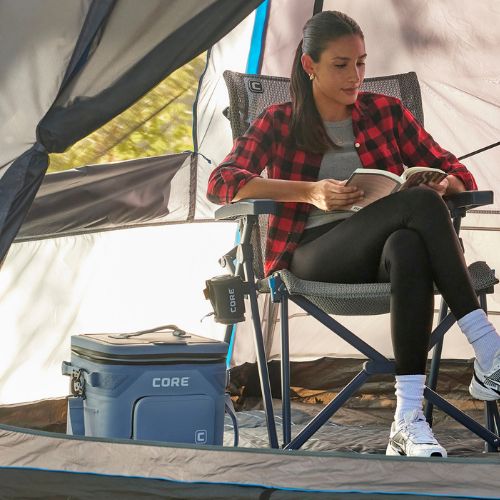 A woman is reading a book in a camping chair next to a CORE 36 can cooler inside a tent.
