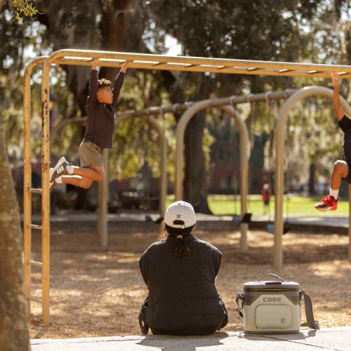 A person sitting on the ground with a cooler next to them, watching children play on monkey bars in a park.