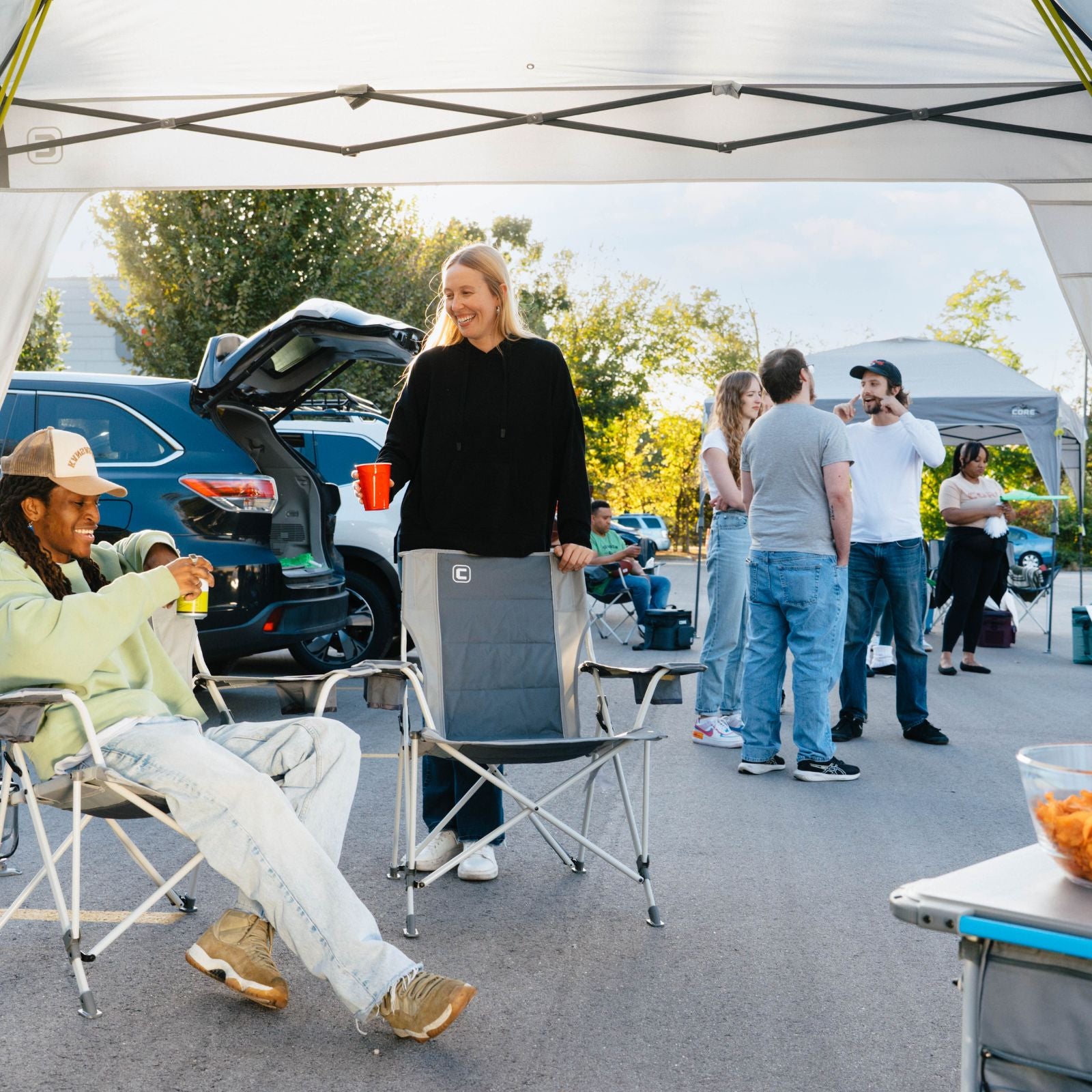 A lifestyle scene at an outdoor campsite or tailgate under a white canopy tent, showing people relaxing in folding chairs. In the foreground, a woman in a black top and jeans stands holding a gray folding chair, while another person sits in a light green chair to the left.