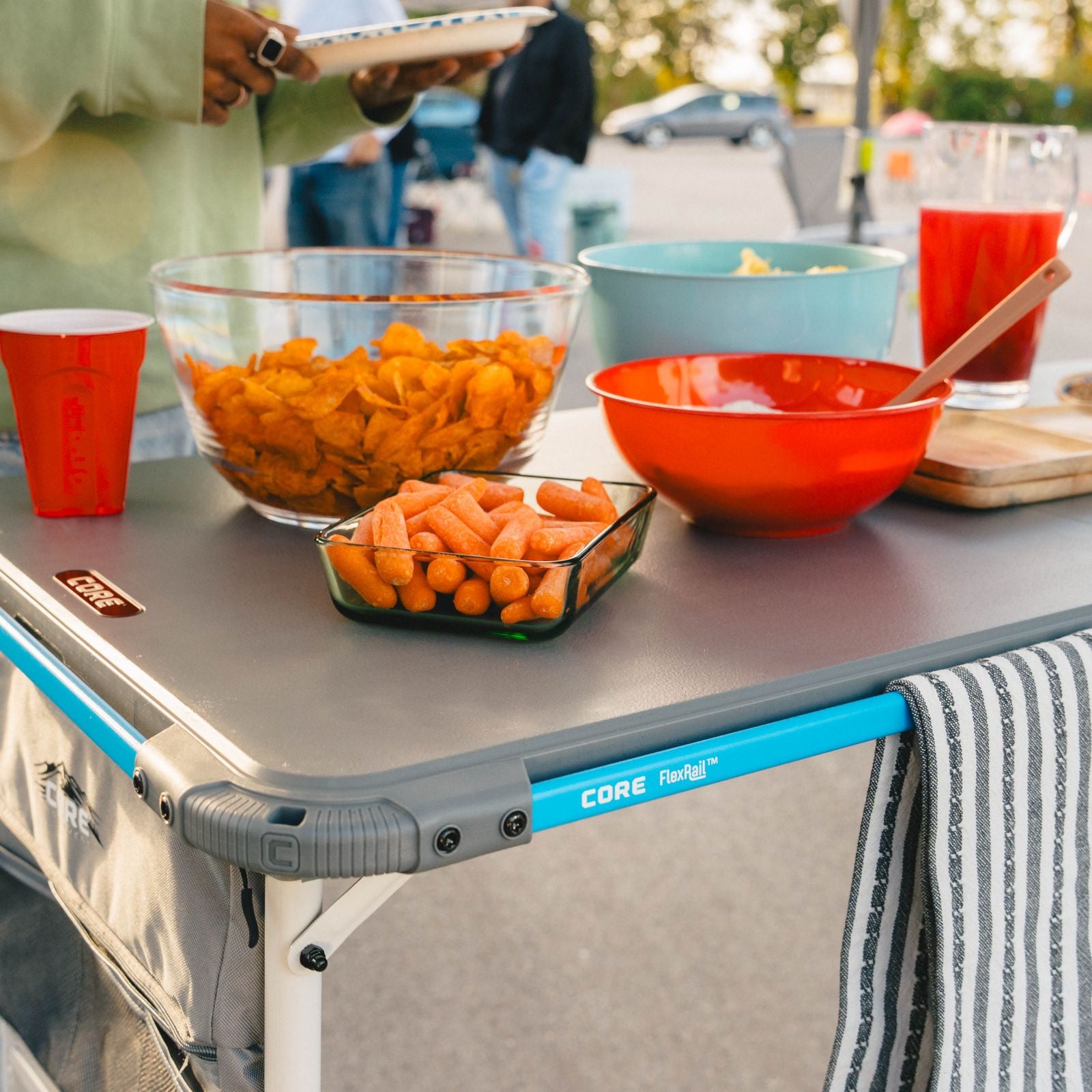 A portable folding table set outdoors with snacks and cups on top. The table has a gray top, bright blue frame accents, and gray corner protectors with the brand name CORE FlexRig visible.