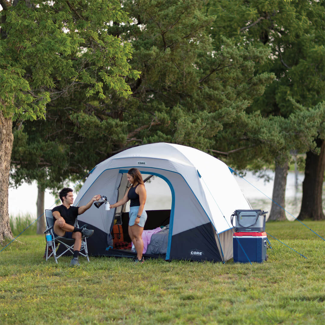 Two people camping outdoors with a CORE 6 person lighted dome tent, chairs, and cooler in a park setting.