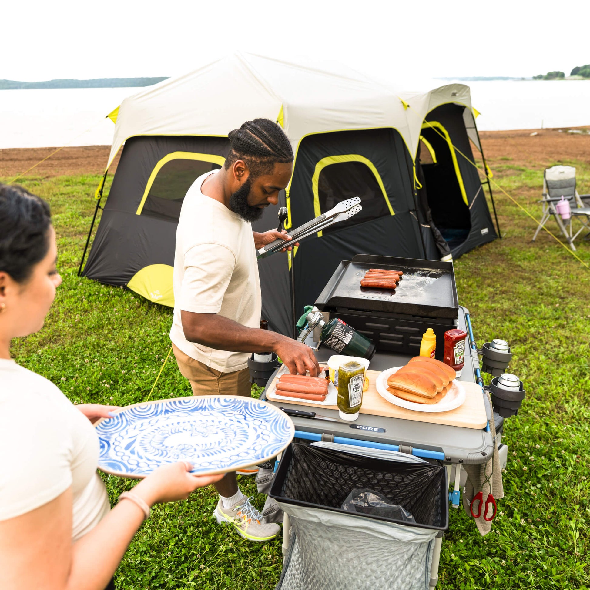 Campers preparing food at CORE outdoor kitchen table with cup holder accessory attached, keeping drinks steady at campsite.