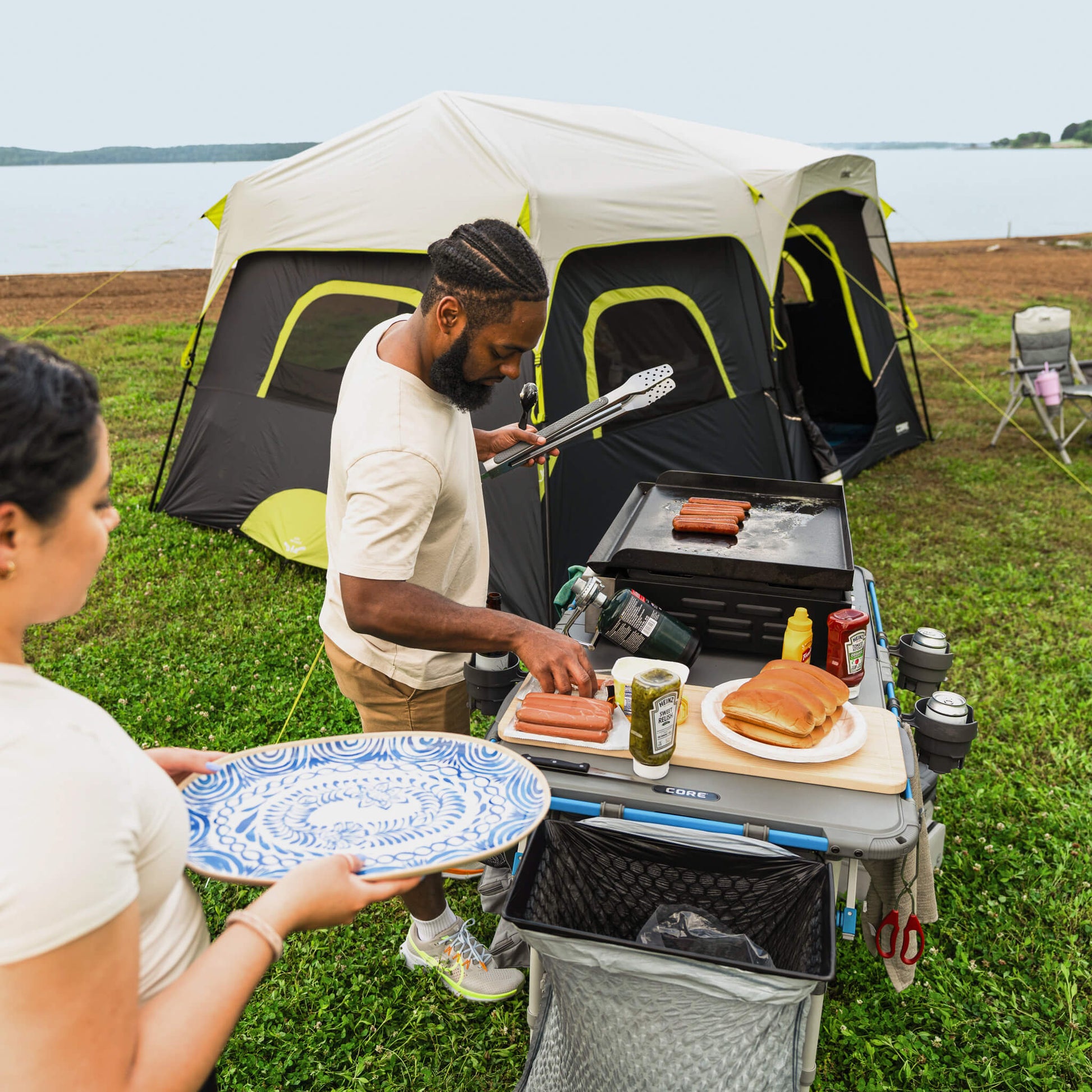 Campers preparing food on the CORE cutting board accessory attached to outdoor kitchen table, with tent setup nearby at campsite.