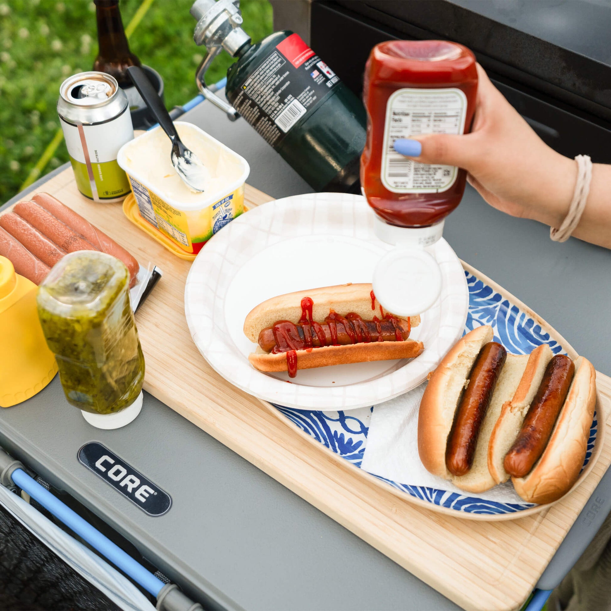 CORE bamboo cutting board accessory in use on 4 foot cook table, holding plates of hot dogs and drinks during outdoor campsite meal.
