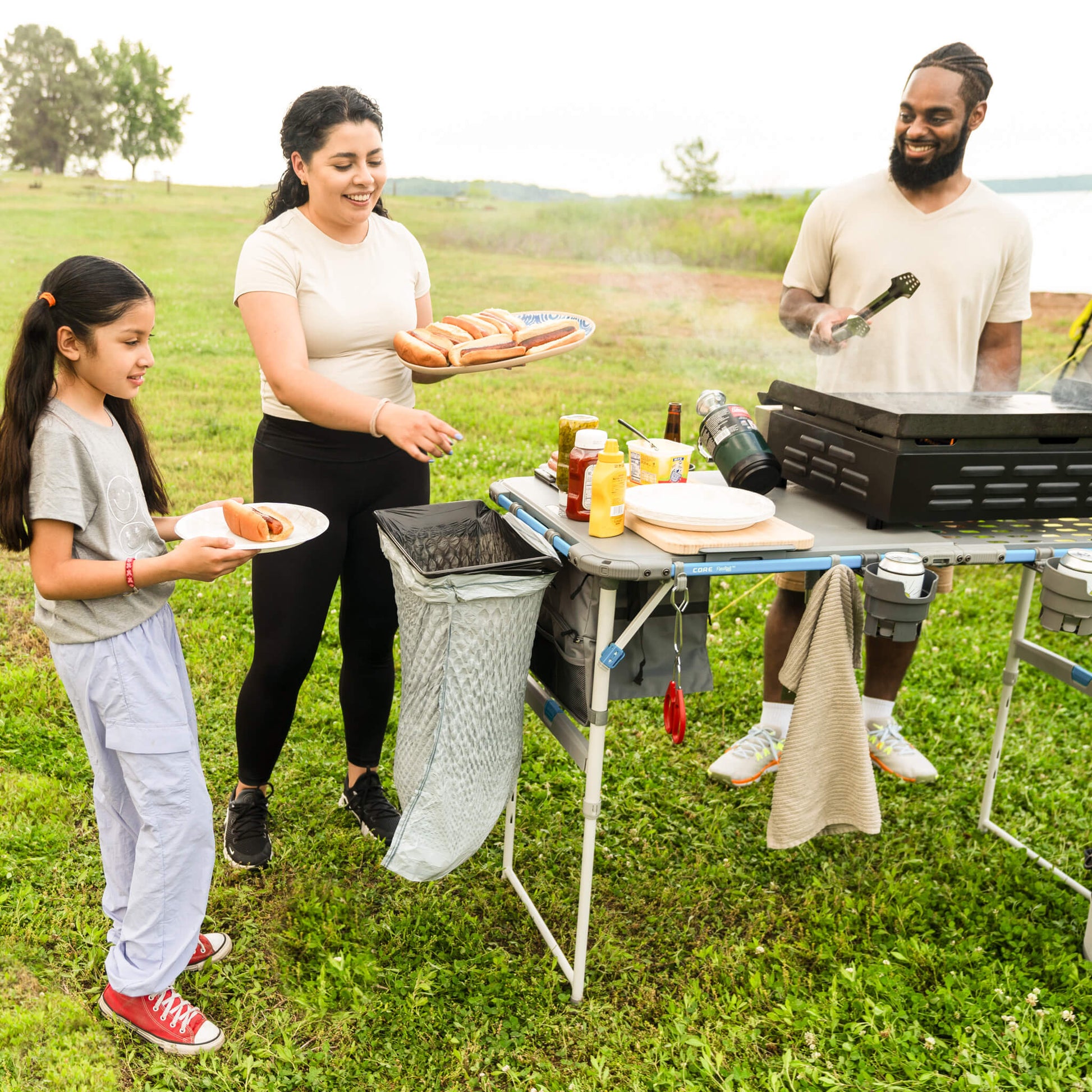 Family preparing food on CORE cutting board accessory attached to 4 foot cook table, enjoying outdoor cooking at campsite.