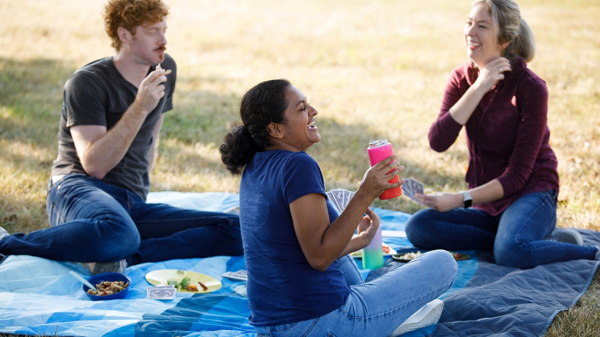 Friends sitting on a picnic blanket outdoors with CORE camping gear, sharing food and drinks on a sunny day.