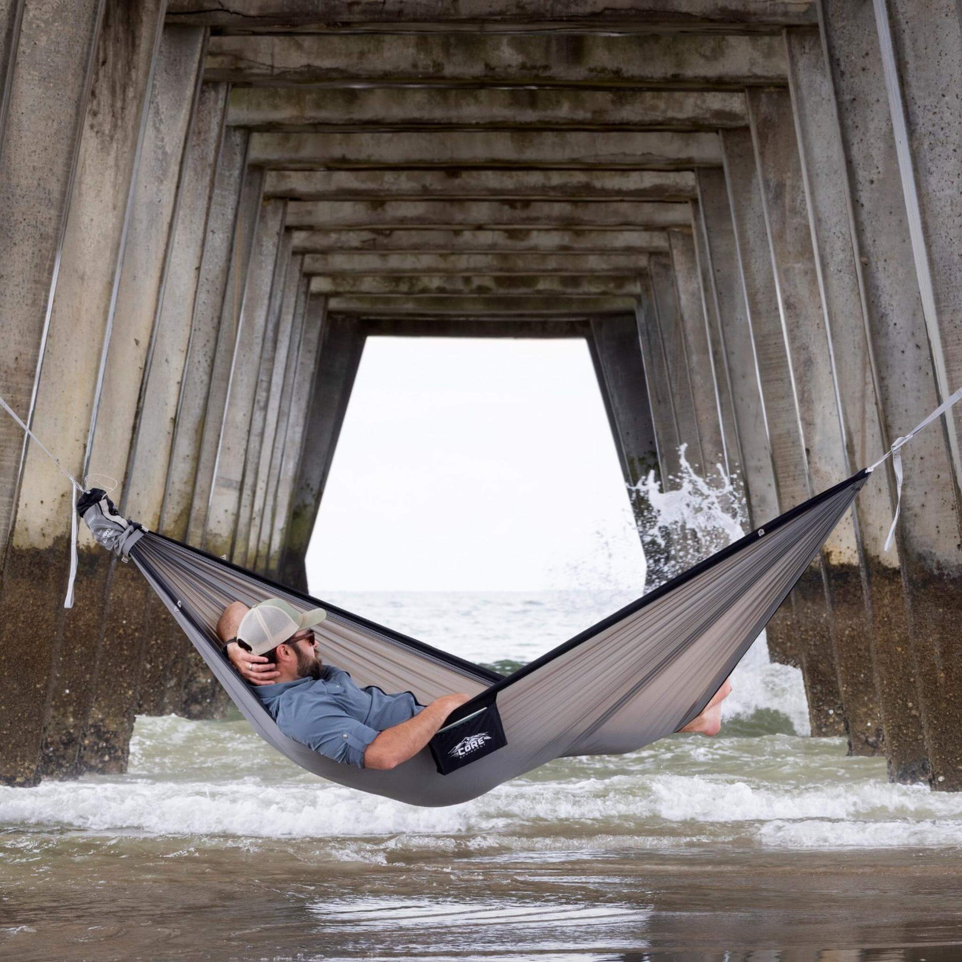 Person resting in a CORE double hammock tied under a pier by the ocean.