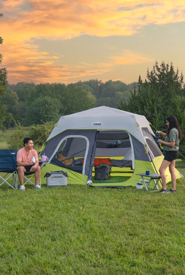 Two people camping outdoors with a tent and picnic setup against a sunset sky.