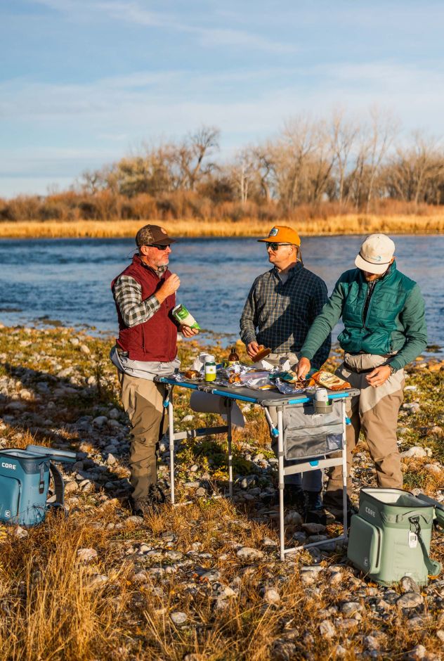 Three men preparing food outdoors by a body of water with fishing gear and coolers.