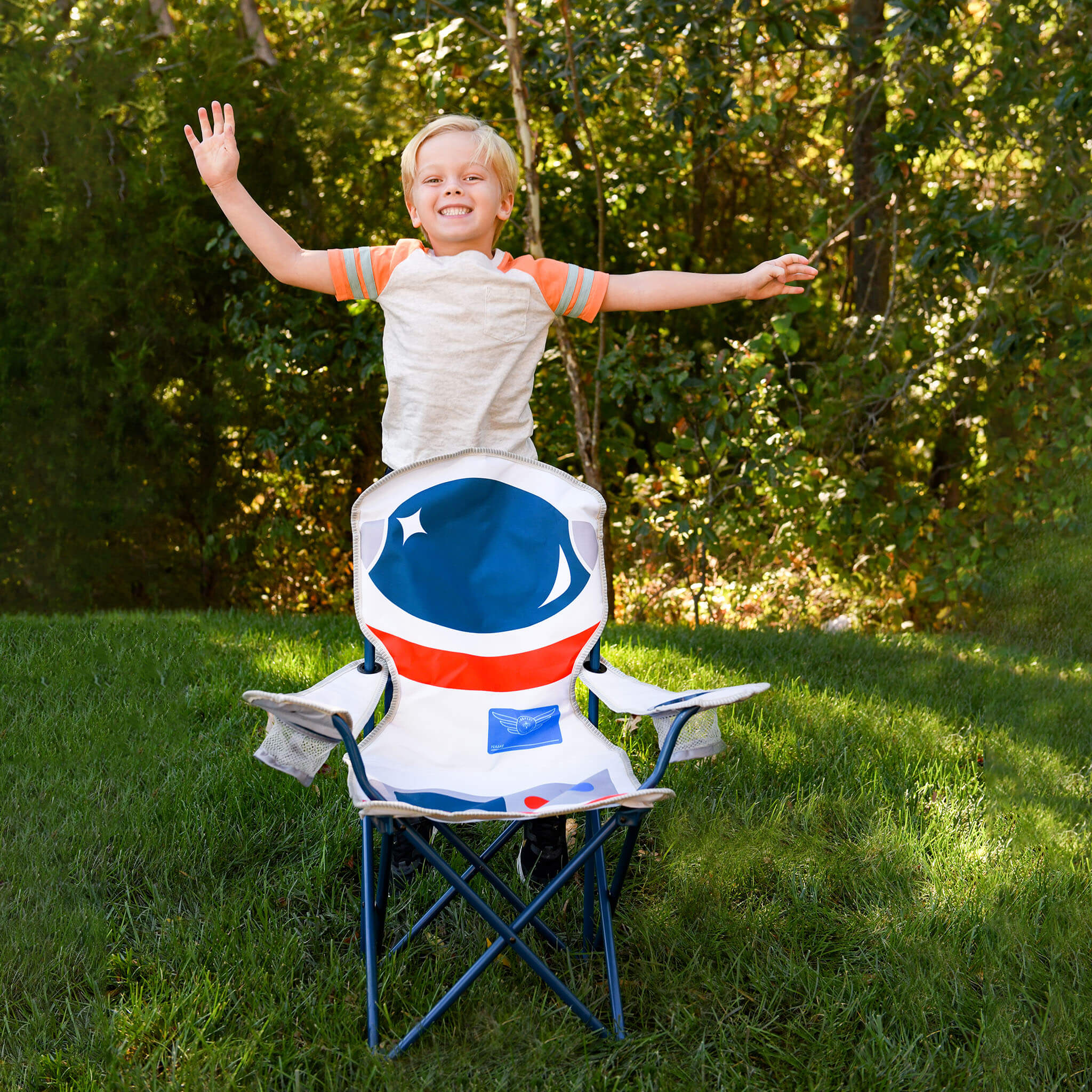 A child stands behind a folding camp chair in a grassy backyard, wearing a white astronaut costume with blue and orange accents. The chair features a large astronaut helmet backrest design in blue, white, and red.