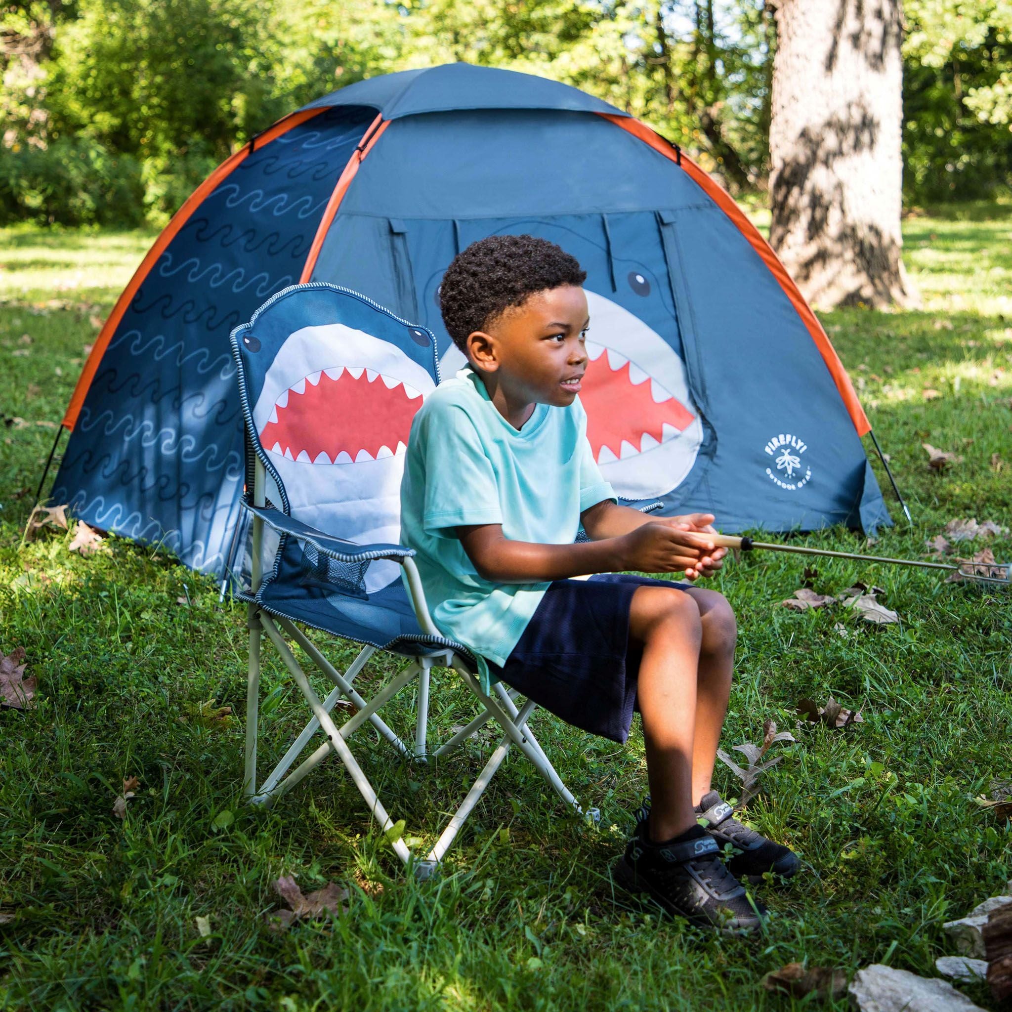 A young boy sits in a blue folding camp chair in a grassy outdoor setting, holding a fishing rod in front of a blue and orange fishing tent with shark-mouth graphics.