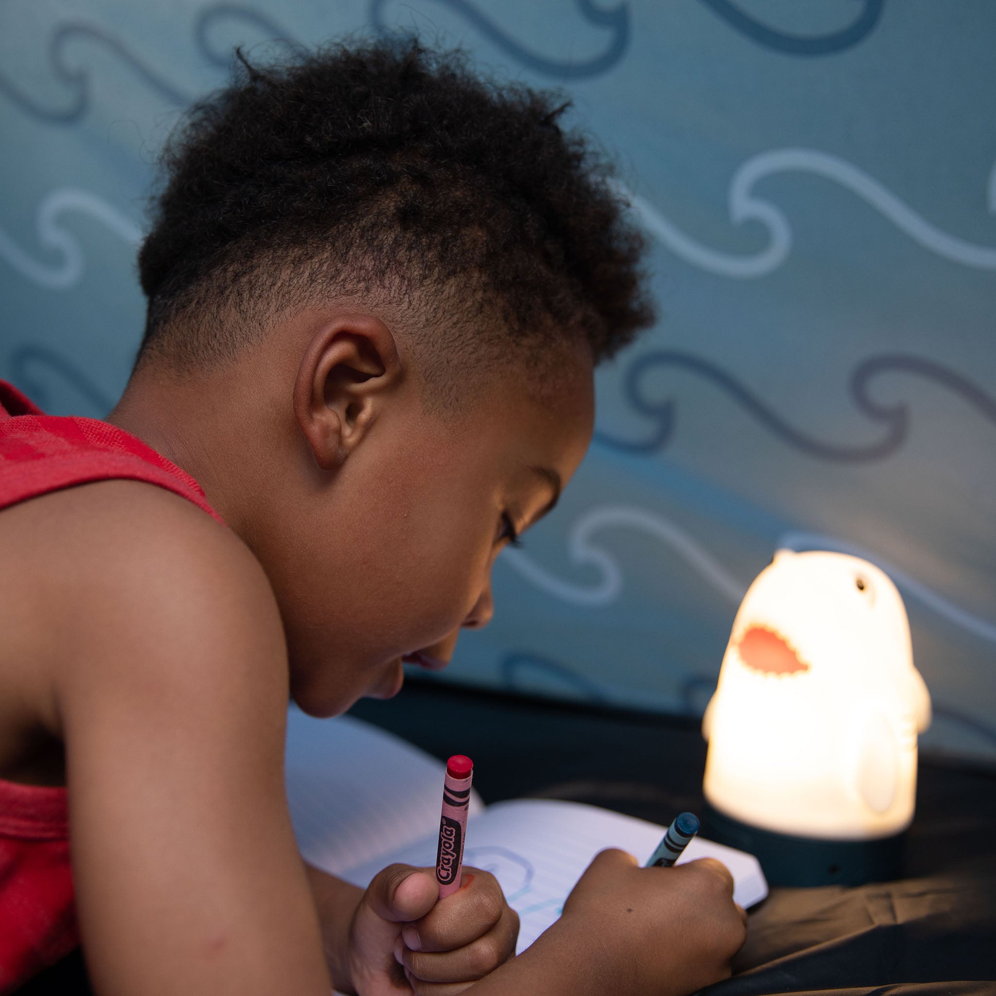 A child drawing with a crayon next to a glowing lamp shaped like a shark.
