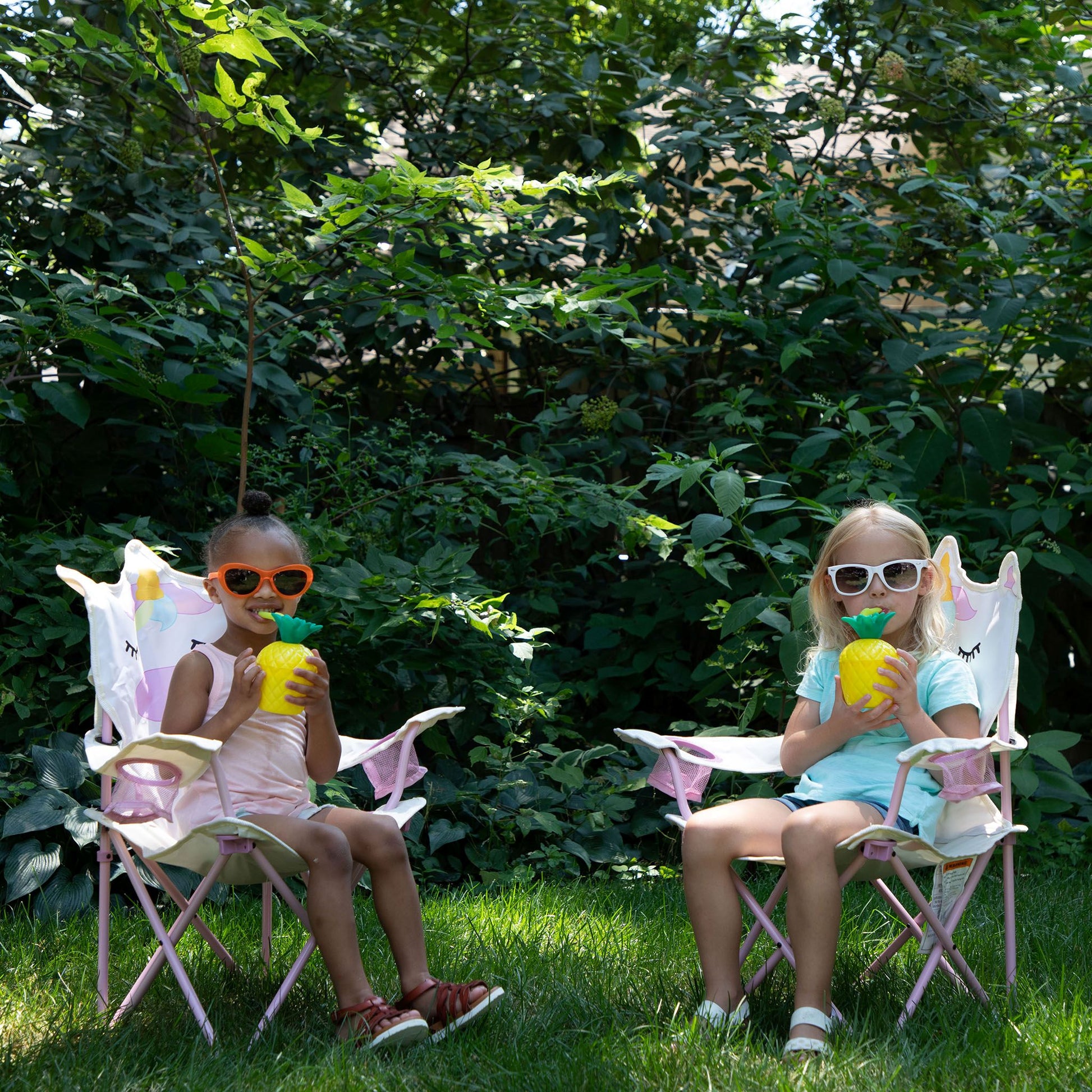Two children sitting in white and pink folding camp chairs on a grassy lawn, each holding a yellow pineapple-shaped drink cup, with a lush green garden background.