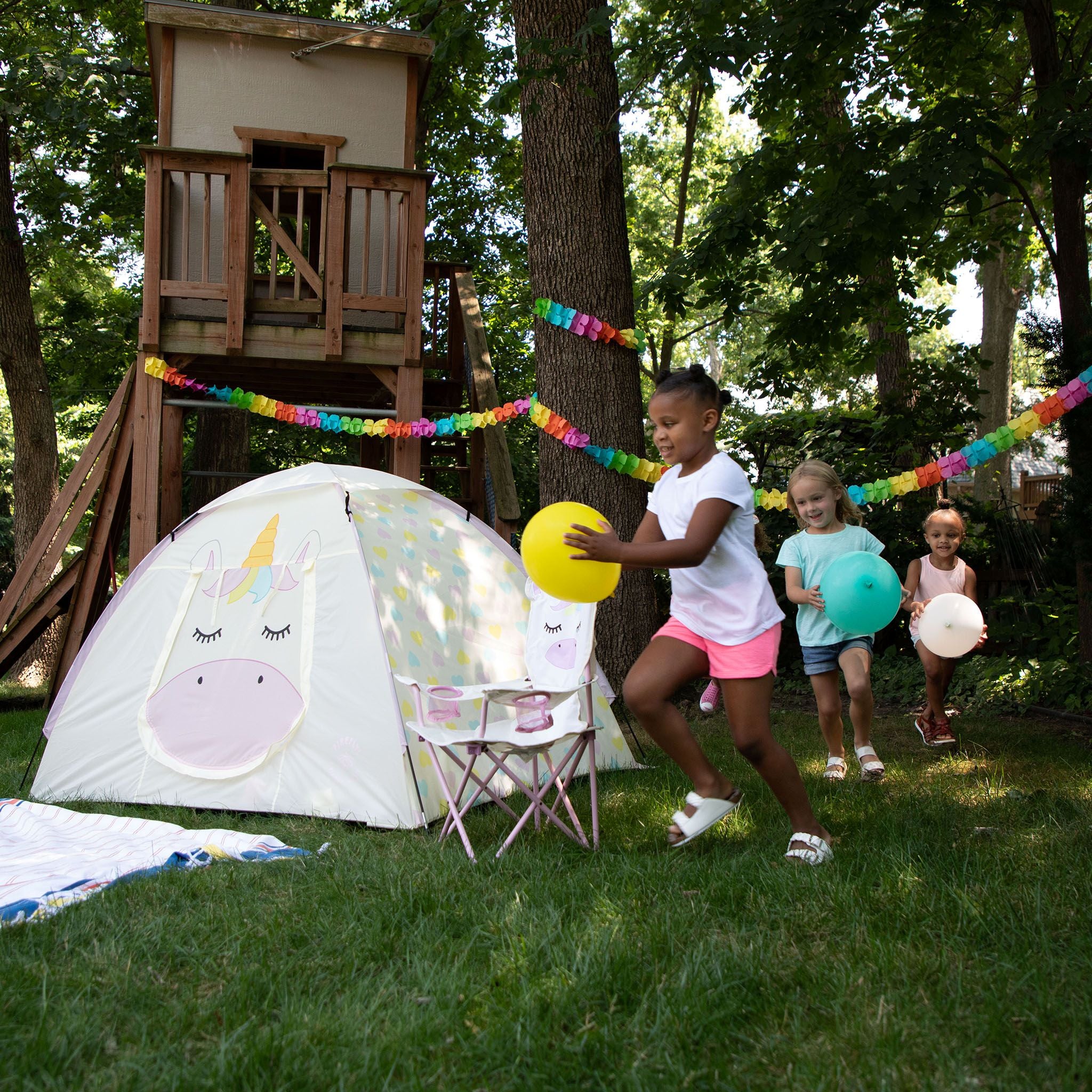 A children's outdoor play scene in a backyard with a wooden treehouse, a white unicorn-themed pop-up tent, a small folding table, and three girls running and throwing colorful balls on the grass. Rainbow paper garlands hang between trees in the background.