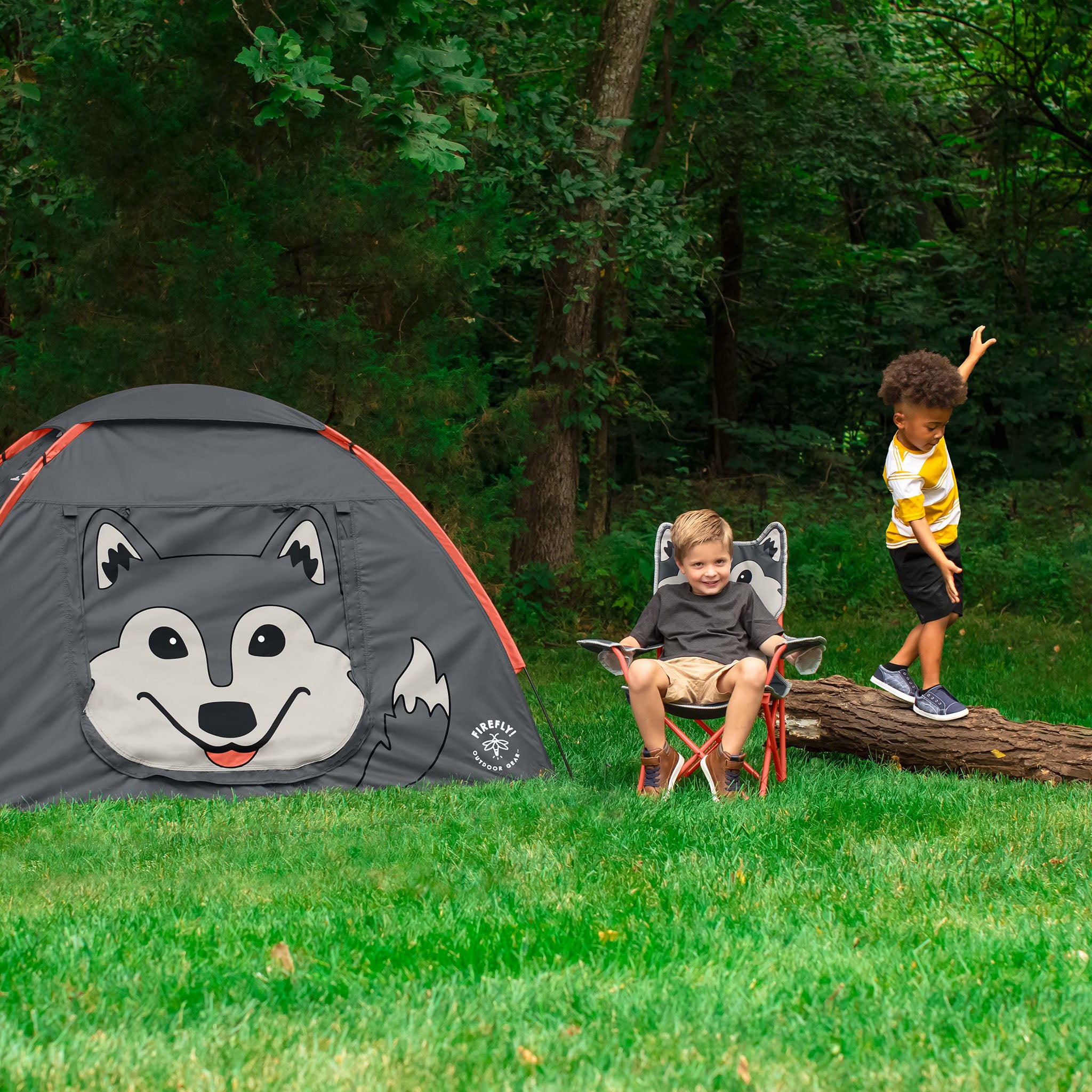 A children's gray camping tent with a large husky face graphic is pitched on green grass in a wooded backyard. Two children are present: one sitting in a matching gray and red camp chair reading a book, and another standing on a log in the background.