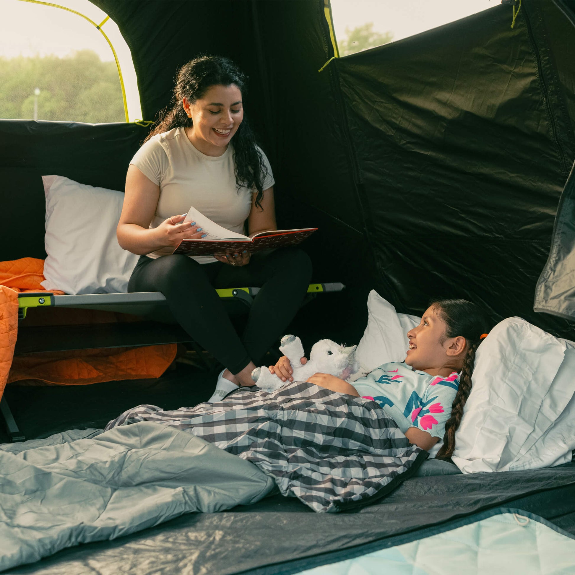 Mother reading to a child sitting on the CORE folding cot inside a tent.