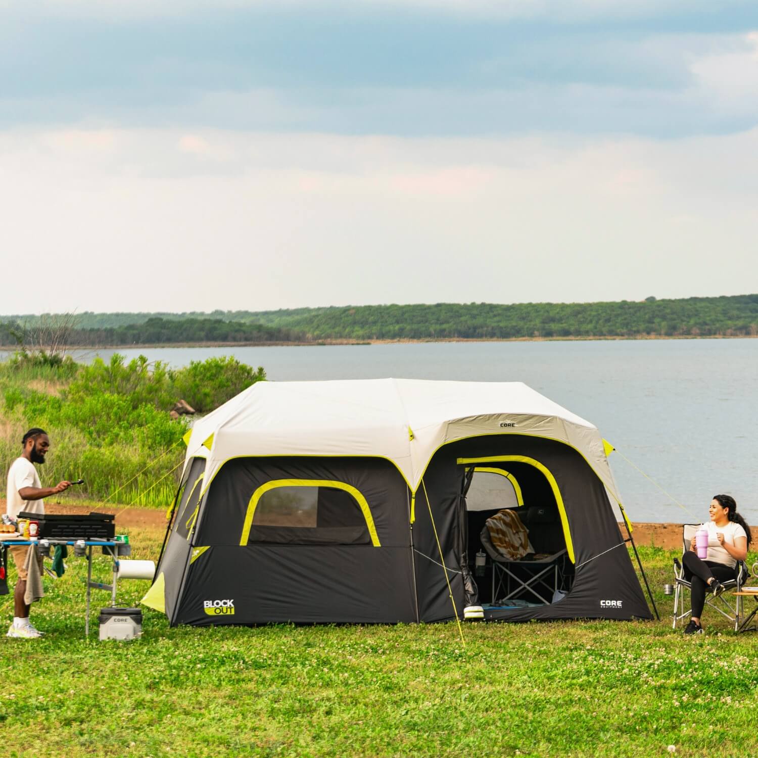 Two people camping beside a lake with a CORE 9 person Block Out tent set up on grassy ground. One person grills food at a picnic table while another relaxes in a camp chair holding a drink. The tent features bright neon green accents and a gray rainfly under a cloudy sky.