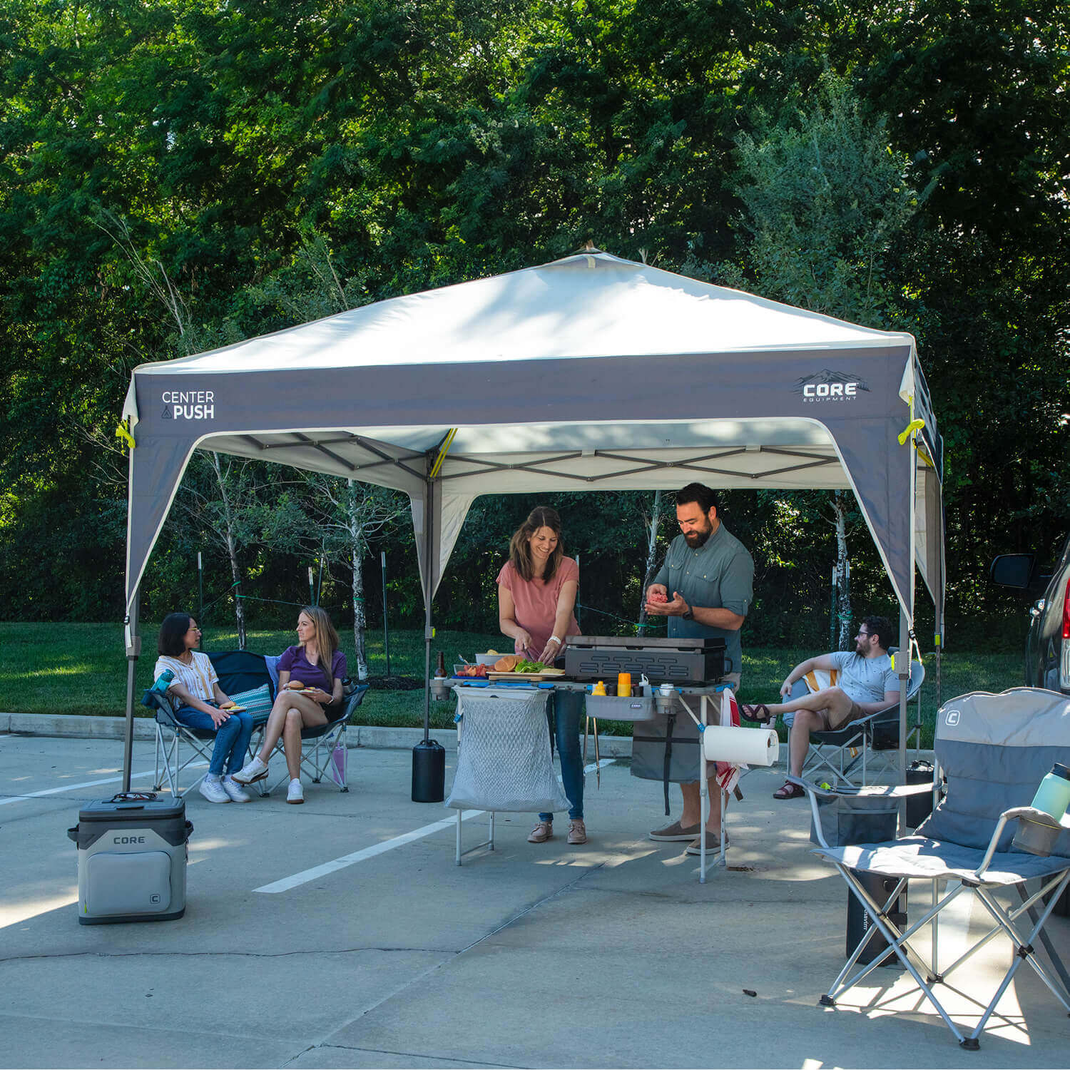 Group of friends relaxing and cooking under a CORE 10x10 Center Push canopy set up in a parking area. Two people prepare food on a table while others sit in camping chairs nearby. A CORE cooler and camping chairs are arranged around the canopy, surrounded by trees and sunshine.