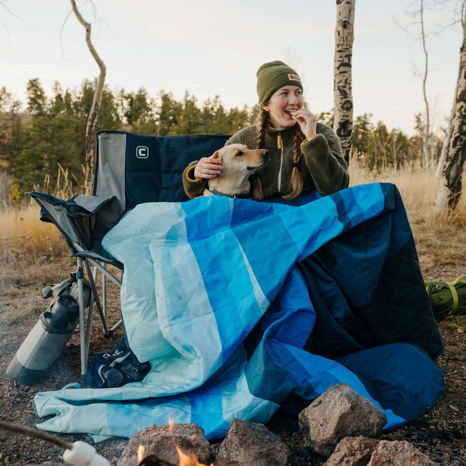 Woman sitting in a CORE camp loveseat chair near a fire, wrapped in a blue outdoor weighted blanket with her dog on her lap. She’s smiling and snacking while enjoying the forest scenery in the background on a cool day.