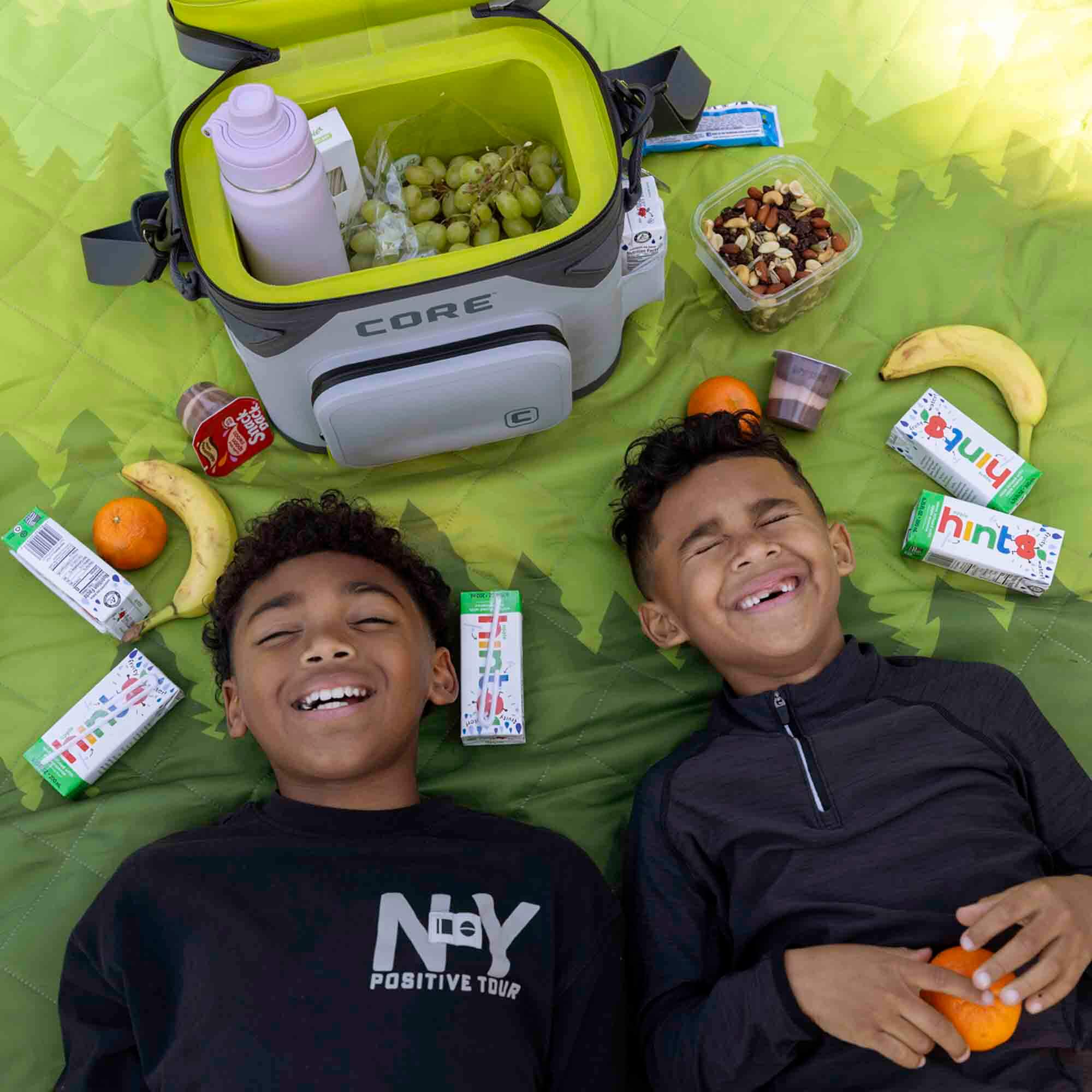 Two children lying on a green CORE weighted blanket with snacks and a CORE Climakeep 12 can cooler.