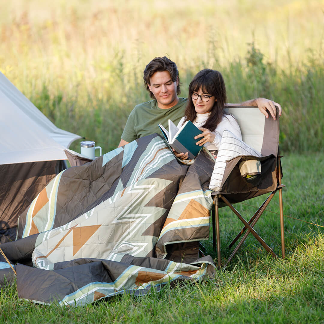 Two people sitting under a patterned weighted blanket on a CORE loveseat in a grassy field with a CORE 6 person lighted dome tent in the background.