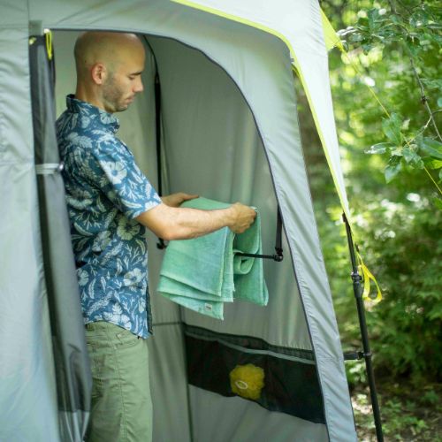 A man is holding a green towel inside a shower tent with trees in the background.