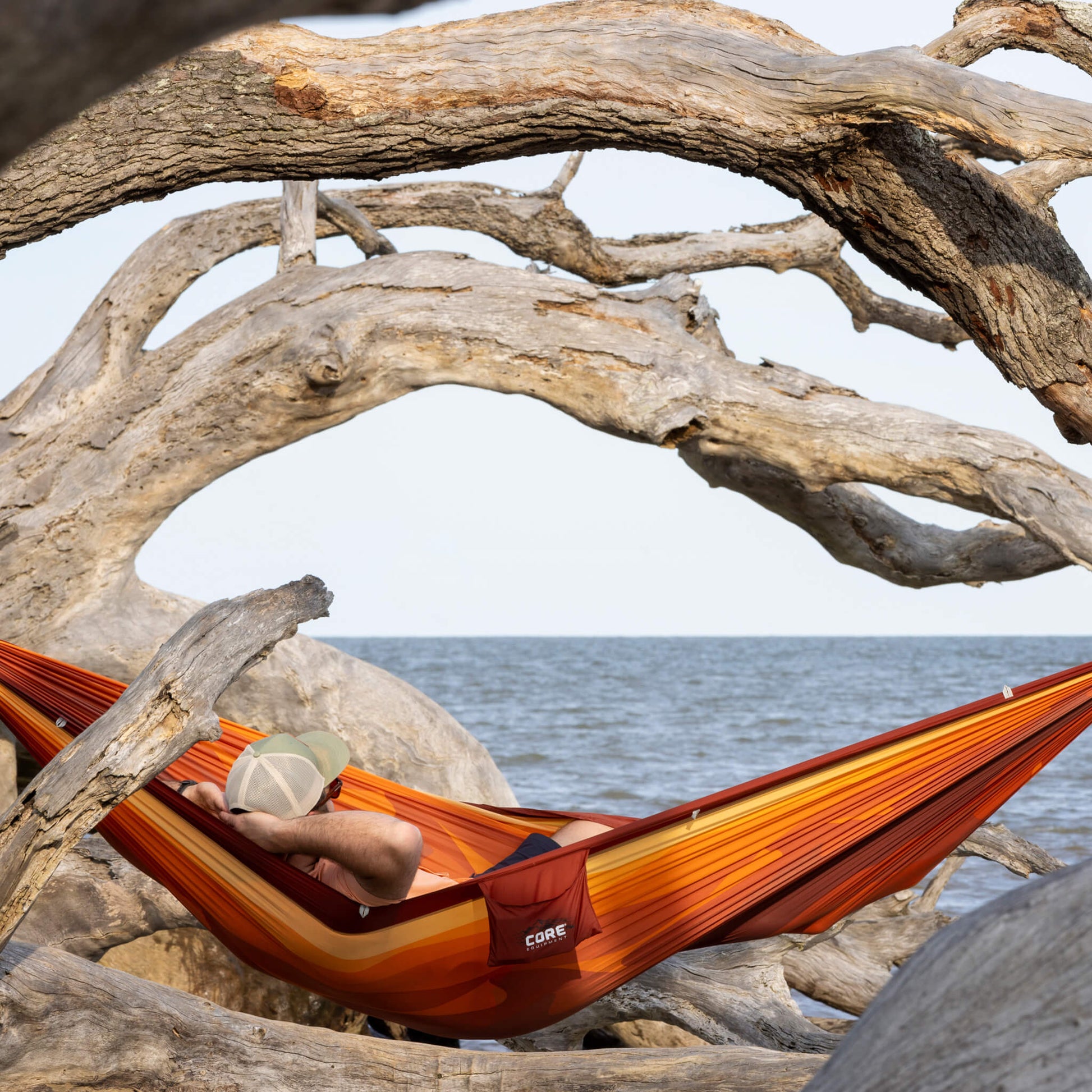 Person relaxing in a CORE single hammock in orange tied between large driftwood trees.