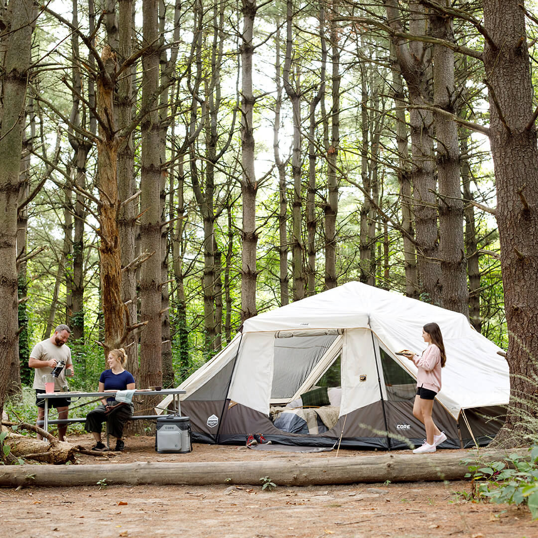 Campers relaxing outside a CORE 6 person lighted instant pyramid tent set up in the forest, enjoying shade and outdoor comfort at the campsite.