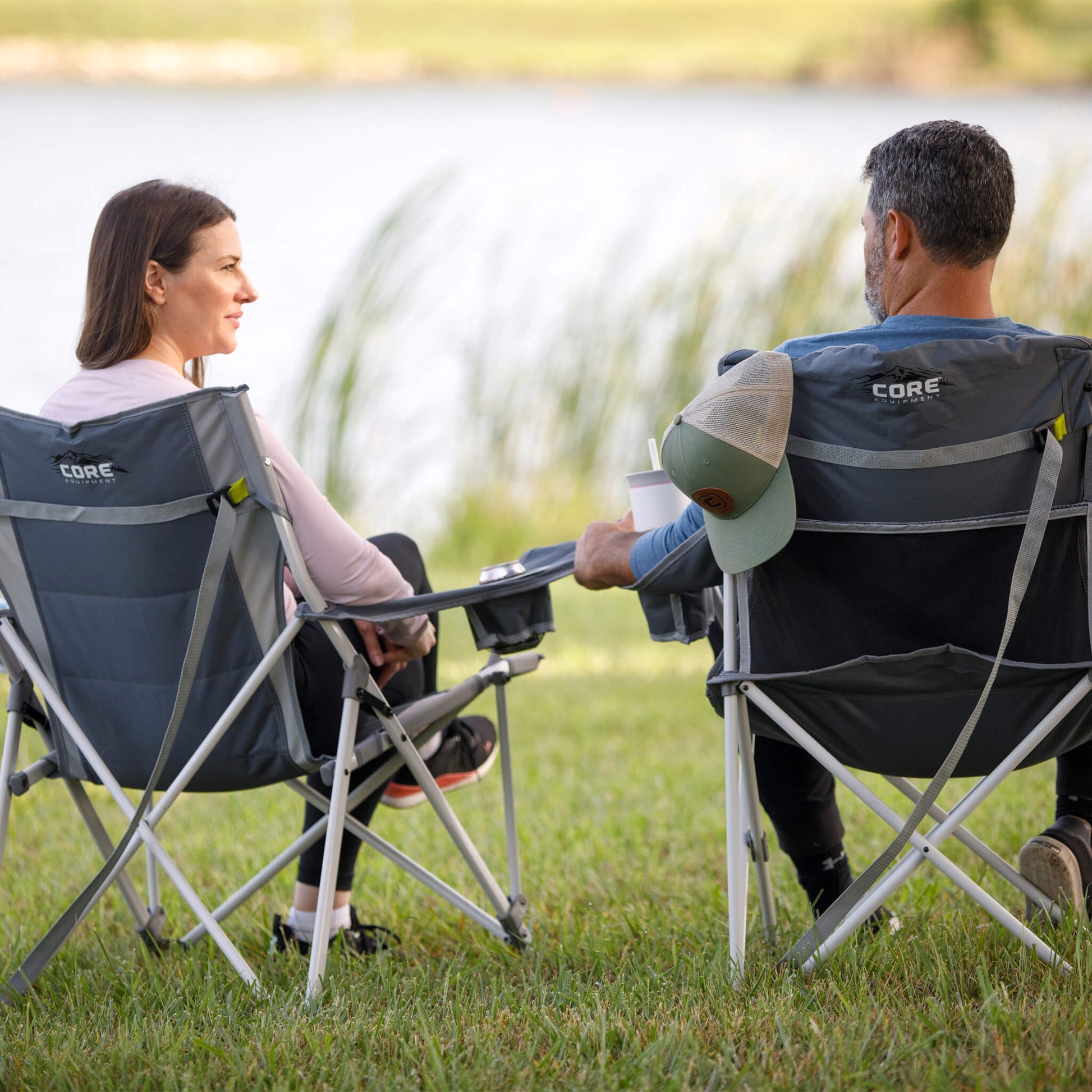 Two campers by the water in a CORE gray tension chair and CORE gray quad chair with built-in cooler.