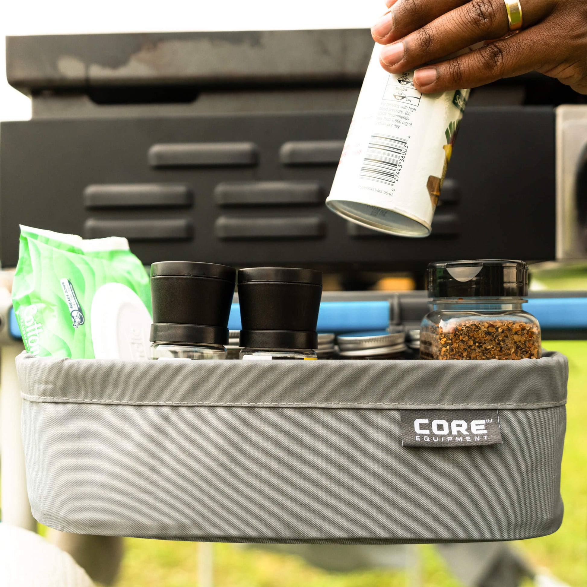A camper using CORE utility basket attached to camp kitchen table, holding spices, seasonings, and cooking essentials outdoors.