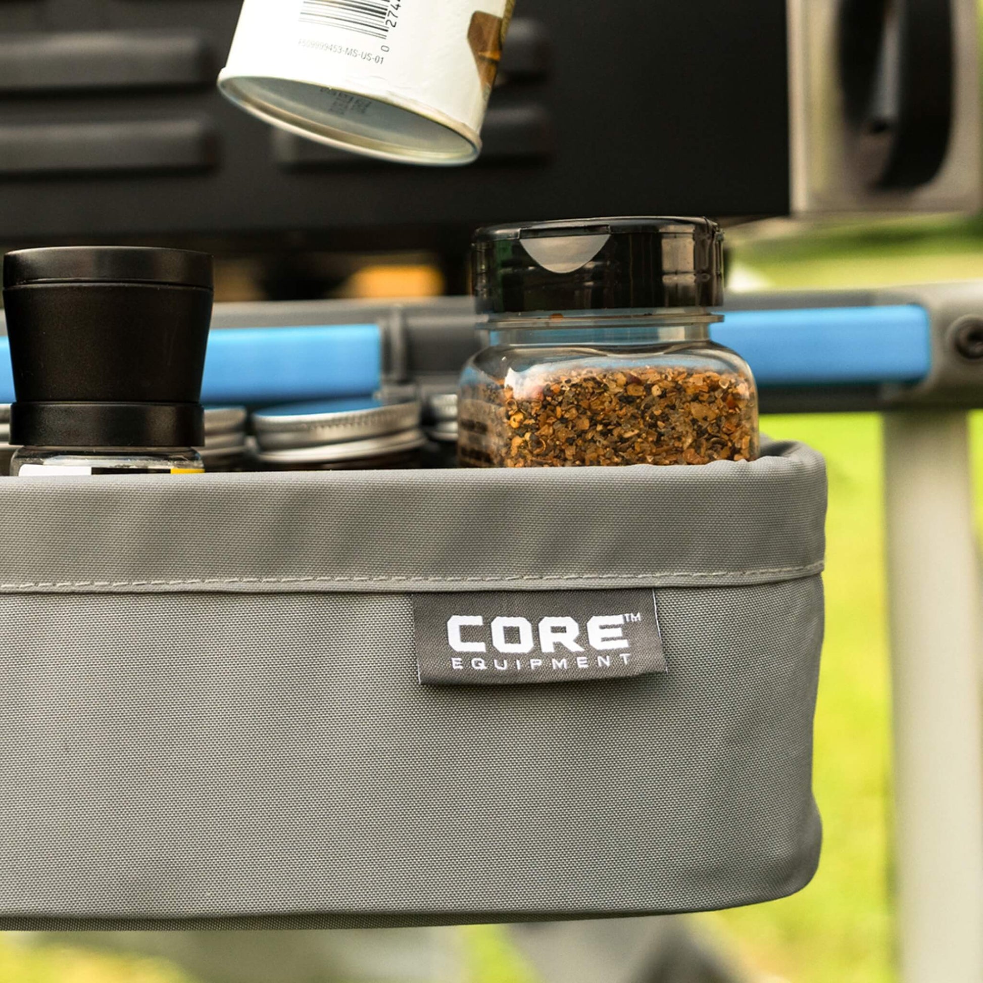 CORE utility basket attached to camp kitchen table, close-up view with jars and spices stored for outdoor meal preparation.