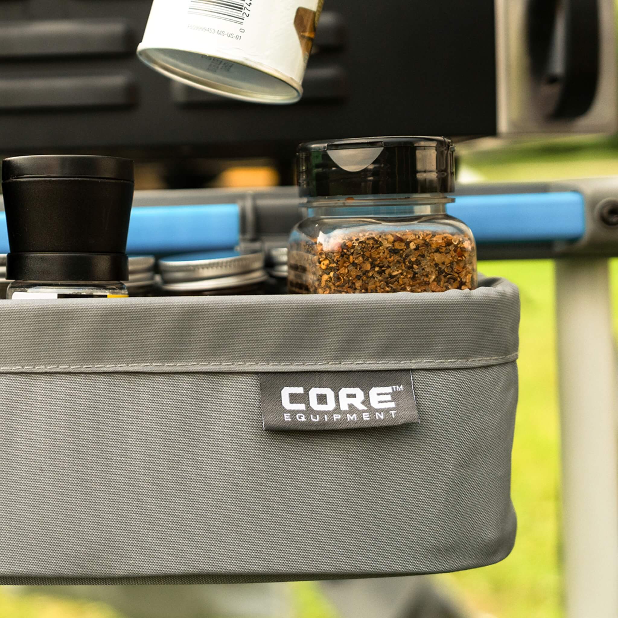 CORE utility basket attached to camp kitchen table, close-up view with jars and spices stored for outdoor meal preparation.