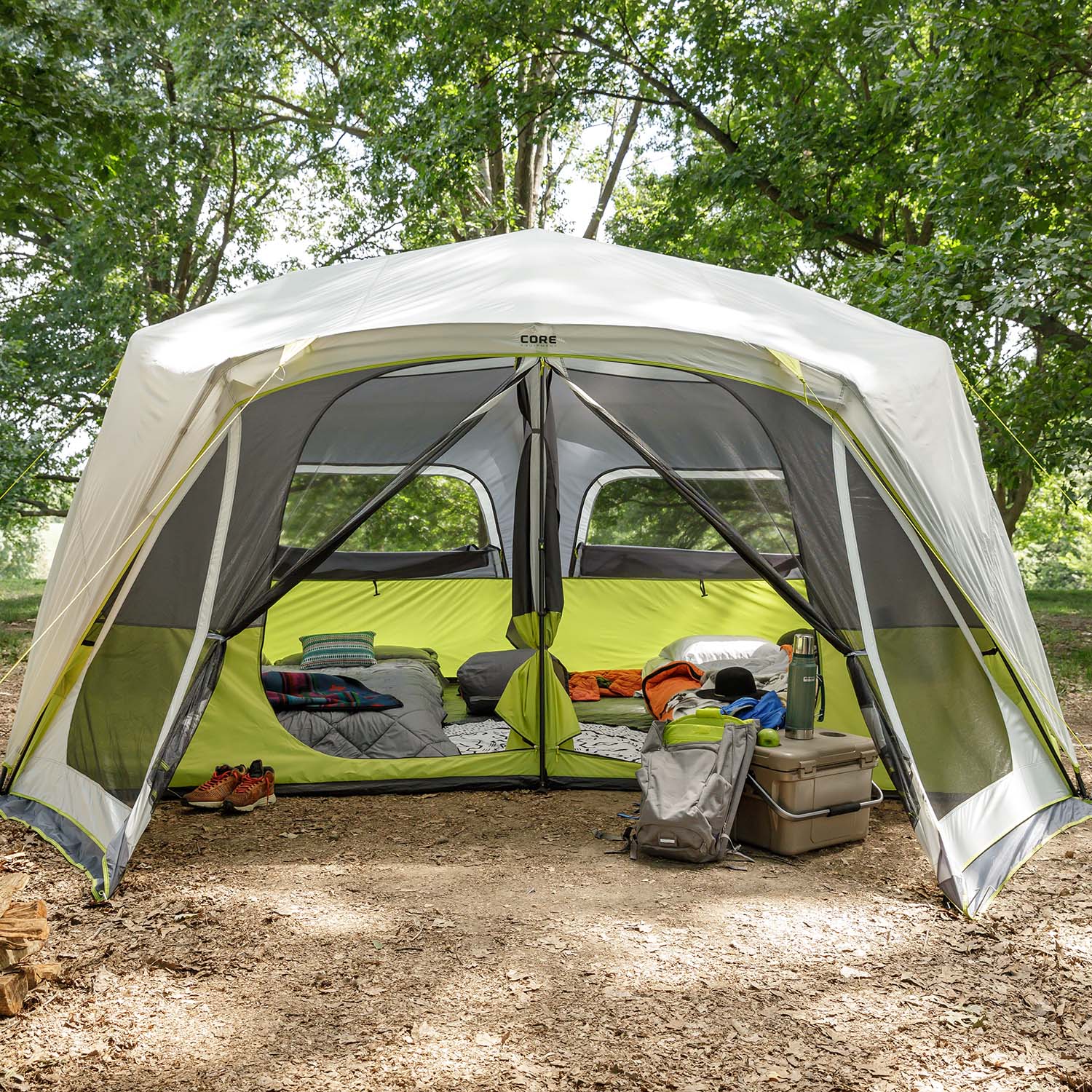 A CORE 10 Person Instant Cabin Tent with Screen Room set up in a wooded area with the front flaps open, revealing two sleeping compartments with bedding. Camping gear, including a backpack, cooler, shoes, and other items, are arranged neatly on the ground outside the tent.