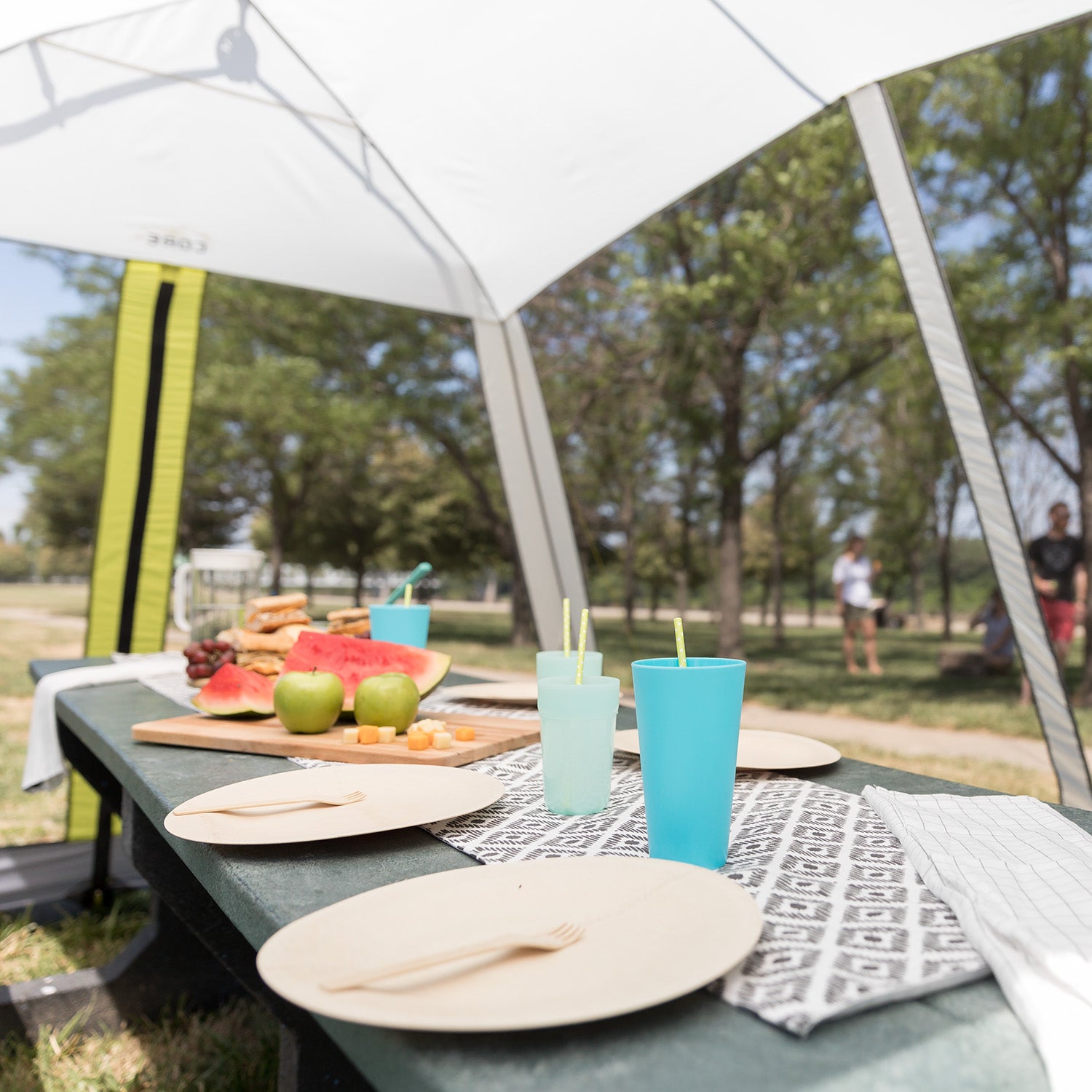 A 12' x 10' Instant Screen House by CORE is neatly set with plates, wooden utensils, and cups. The table features a colorful spread including watermelon, apples, sandwiches, grapes, and other snacks. Trees and people relaxing in a park are visible in the background.
