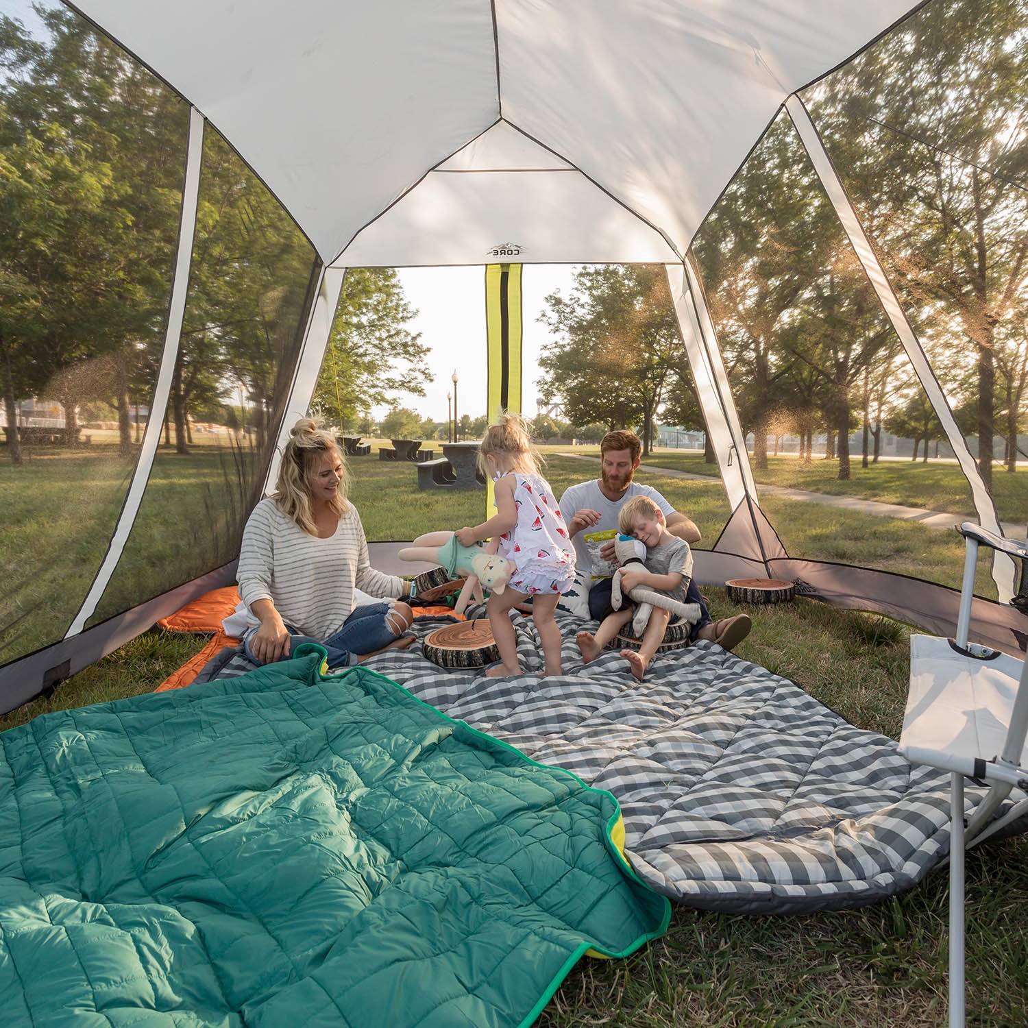 A family of four enjoys time inside a large CORE 12' x 10' Instant Screen House pitched in a grassy area. Parents sit on a checkered mat while one child stands and another crawls. A green sleeping bag and camping gear are visible, and trees can be seen through the mesh walls.