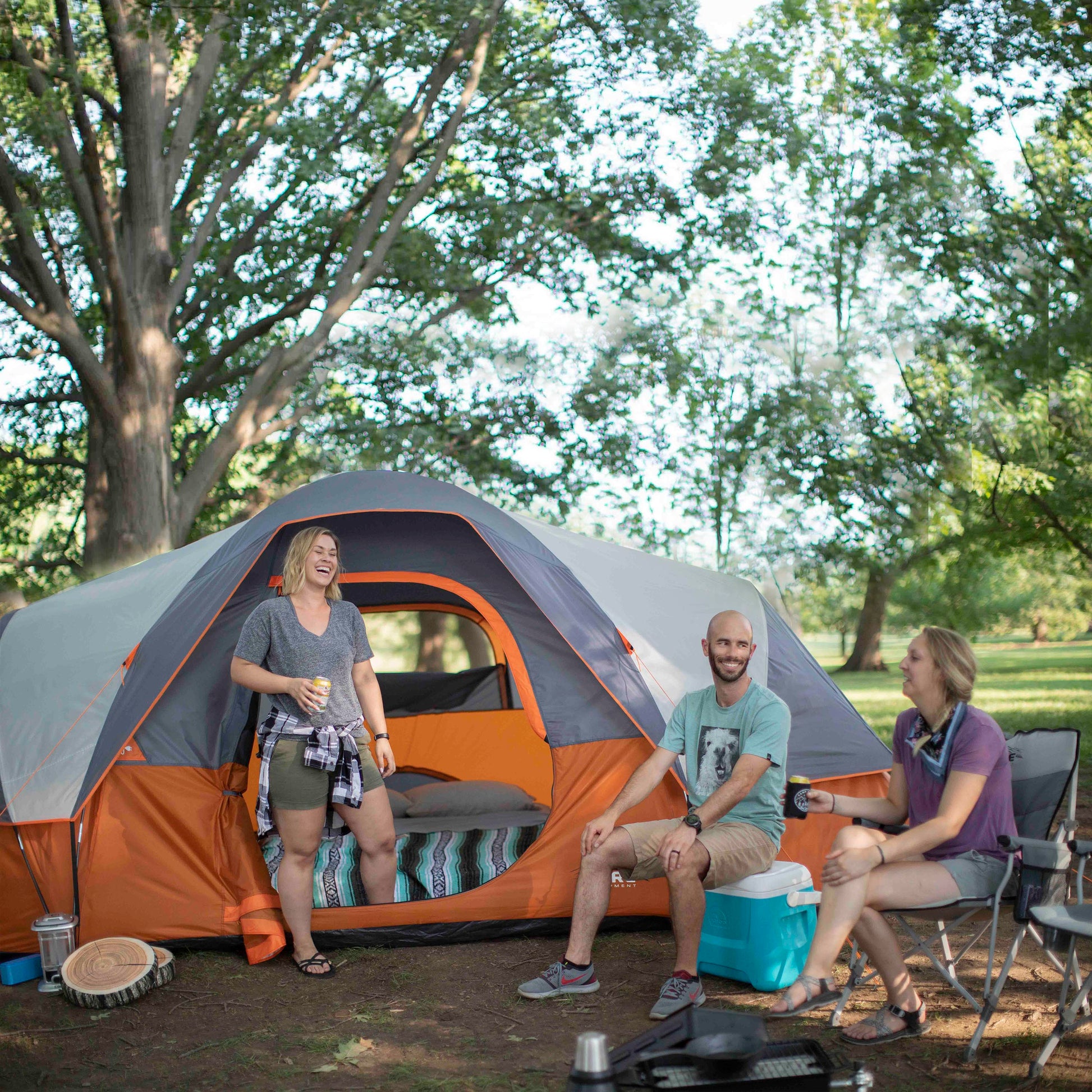 Three people are gathered outside a large orange and gray CORE 9 Person Extended Dome Tent in a wooded area. They are smiling and chatting, enjoying a camping trip. One woman stands by the tent holding a drink, while a man and another woman sit on a CORE chair and blue cooler.