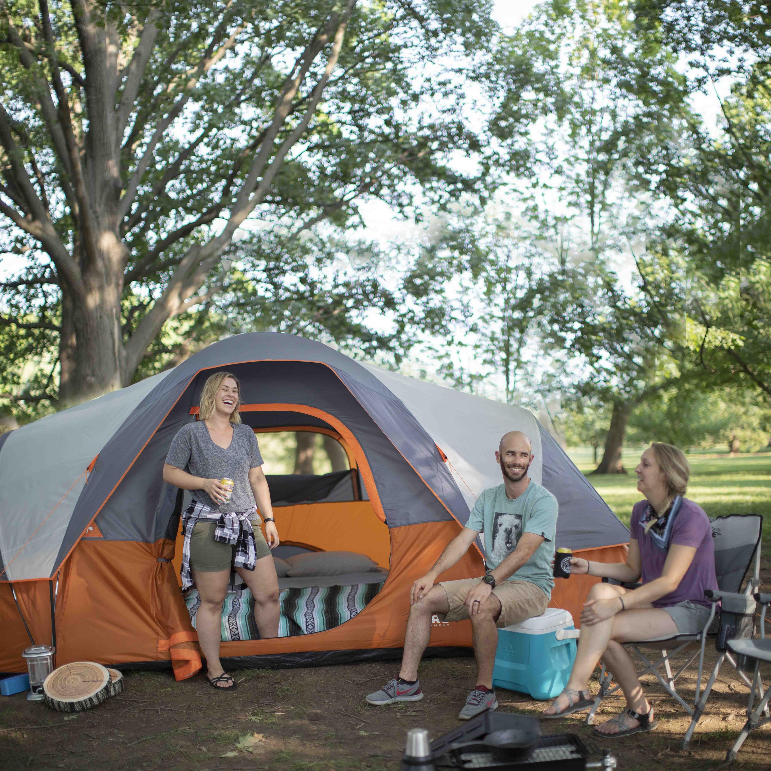 Three people are gathered outside a large orange and gray CORE 9 Person Extended Dome Tent in a wooded area. They are smiling and chatting, enjoying a camping trip. One woman stands by the tent holding a drink, while a man and another woman sit on a CORE chair and blue cooler.