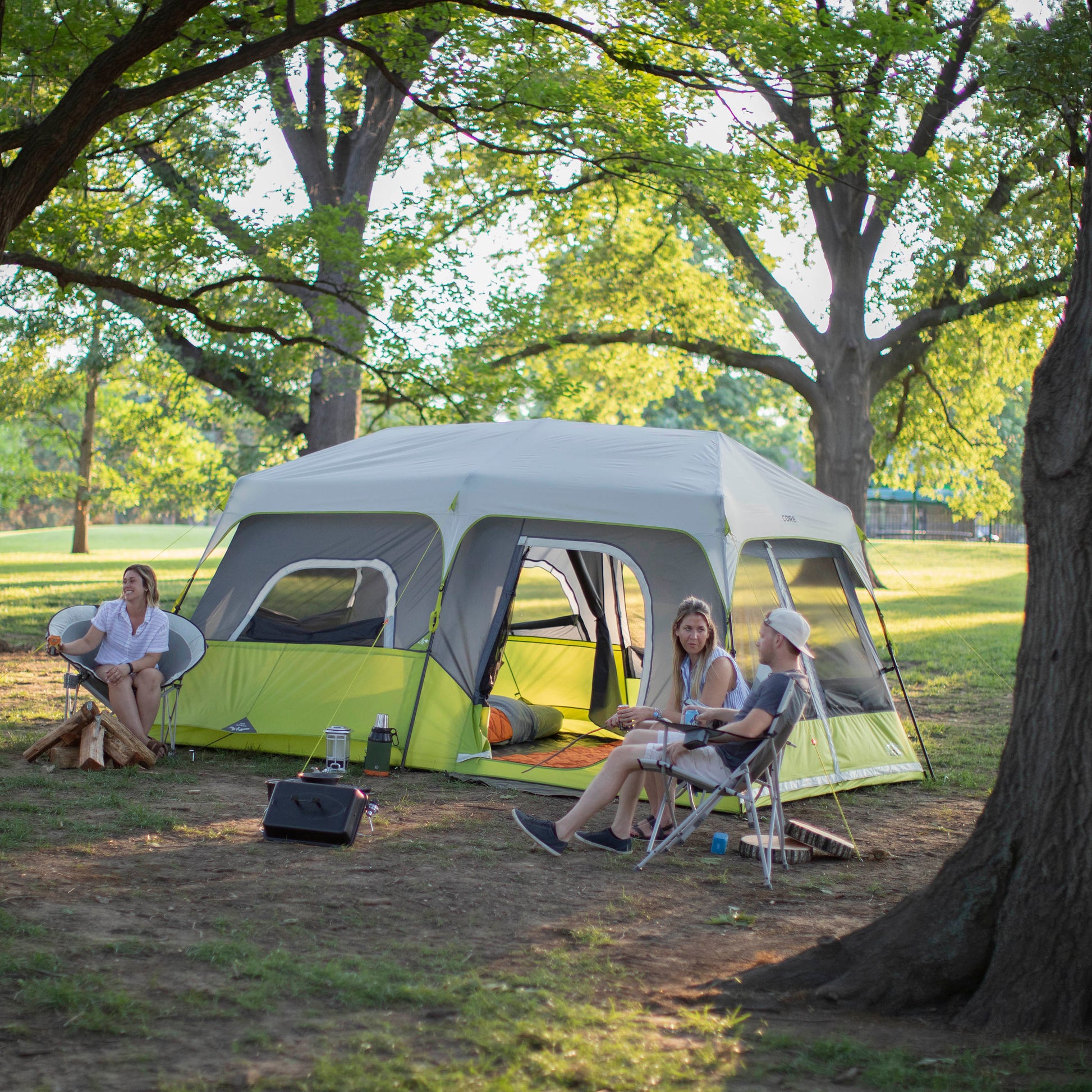 Three people sit outside a large green and gray CORE 9 Person Instant Cabin Tent set up in a grassy park with tall trees. One person is in a folding chair, another on a stool, and the third on the CORE round chair. Camping gear, including chairs and a lantern, is scattered around. The scene is sunny and relaxed.