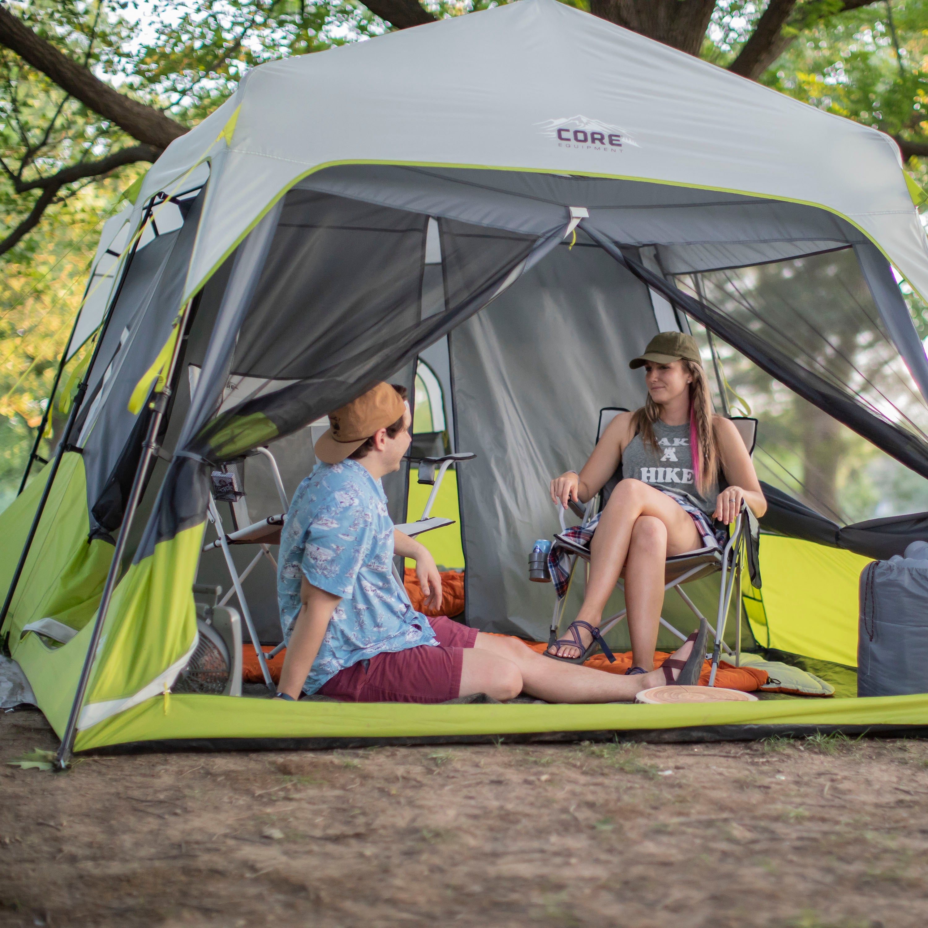 Two people sitting inside a CORE 9 Person Instant Cabin Tent at a campsite. One person is lounging on a mat, wearing a blue shirt and red shorts, while the other sits on a camping chair, wearing a tank top and shorts. Both appear relaxed and are surrounded by outdoor gear and trees.